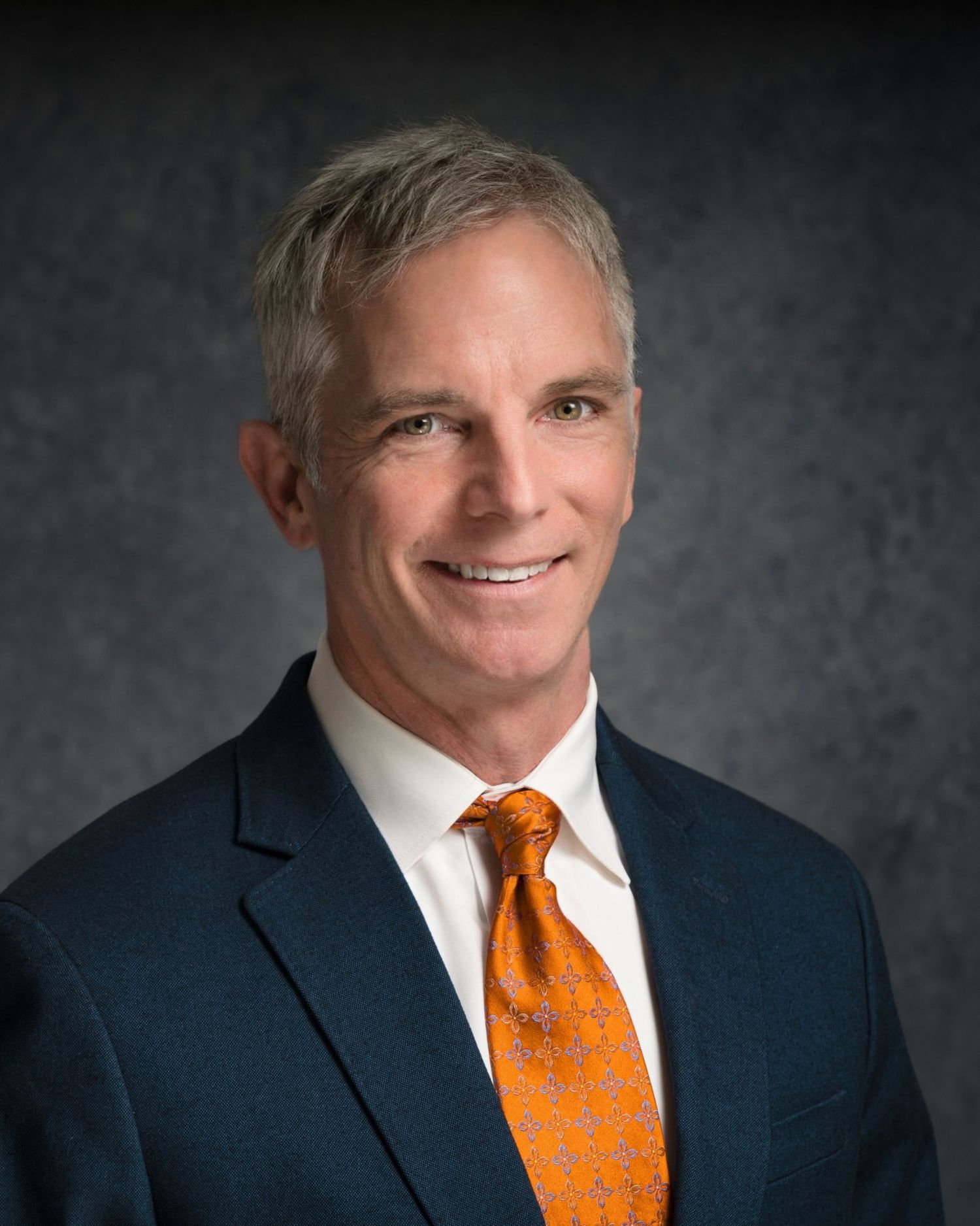 Professional portrait of person in navy suit with orange patterned tie against dark backdrop.