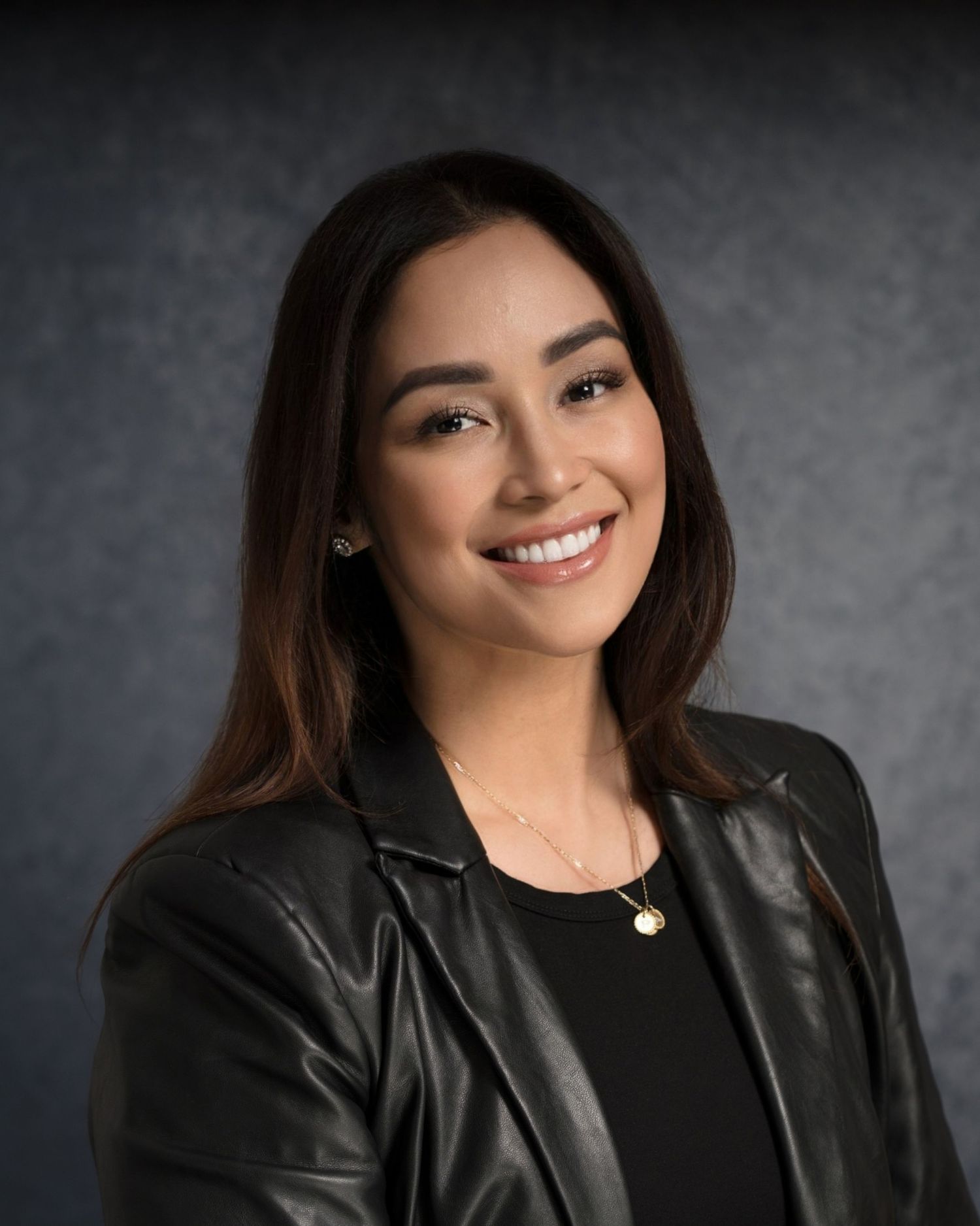 Professional portrait of person in black leather jacket and necklace against dark backdrop.