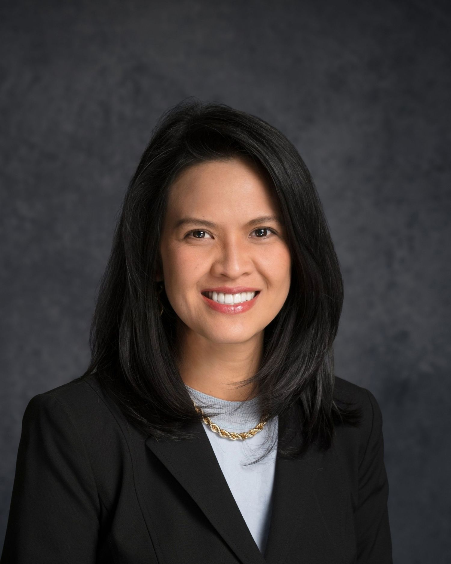 Professional headshot of person in black blazer and light top with layered necklaces.