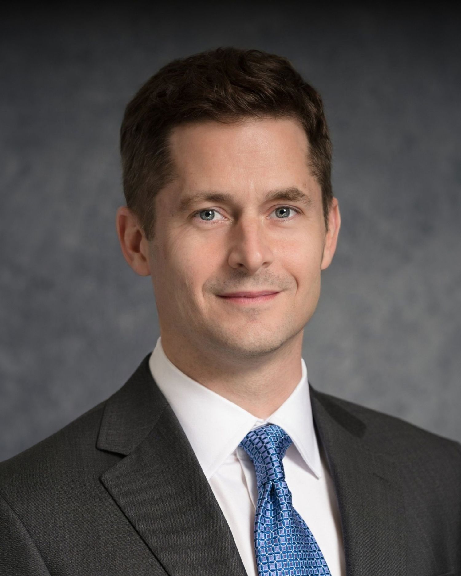 Professional headshot of man in dark suit with blue tie against gray background.