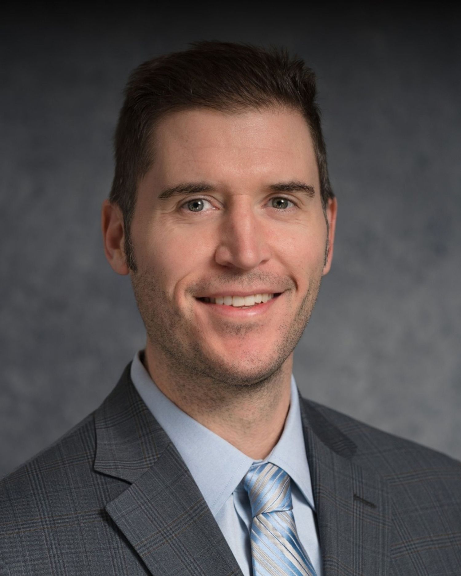 Professional headshot of person in grey suit with striped blue tie against dark background.