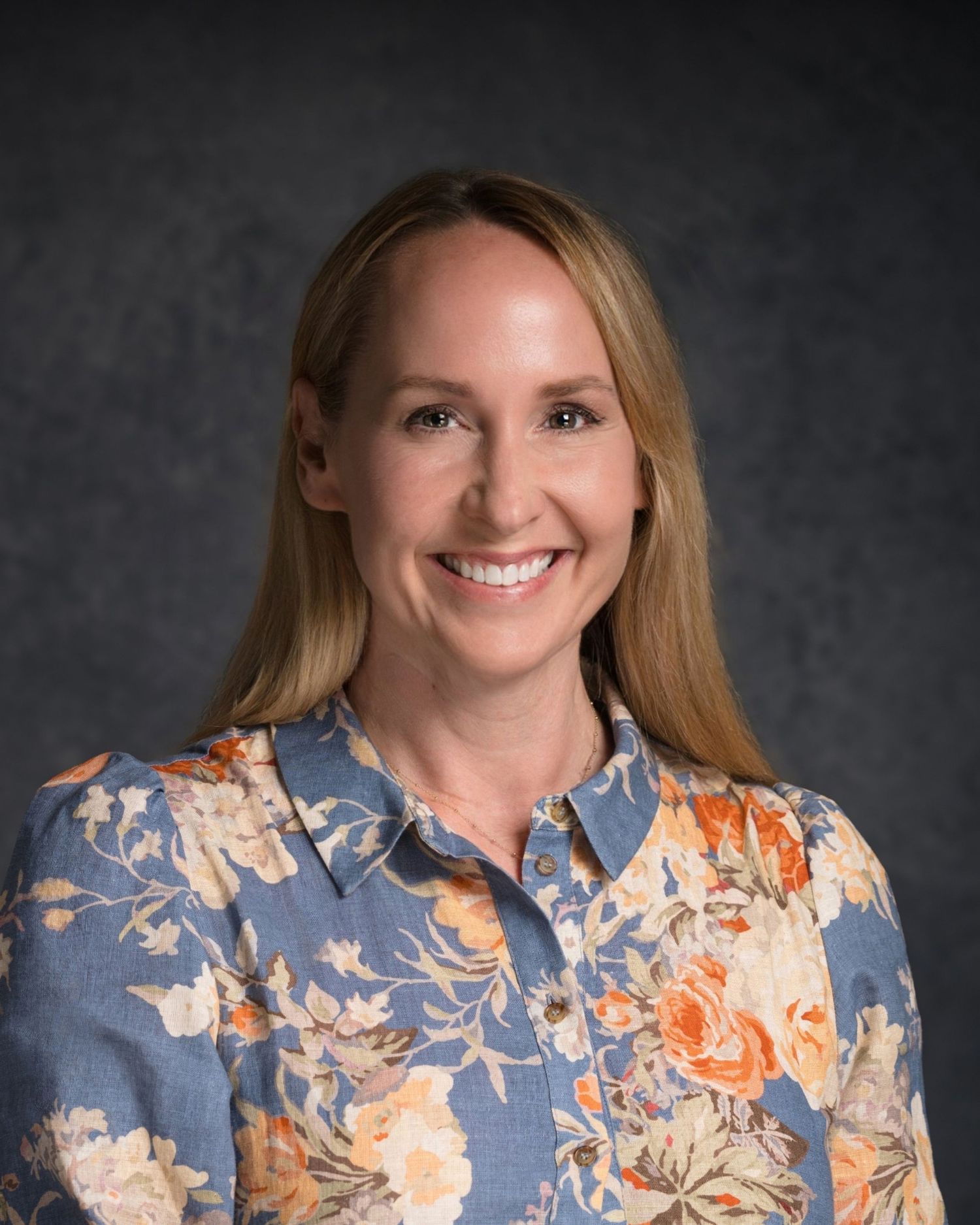Professional portrait of person wearing floral blouse with blonde hair against dark backdrop.