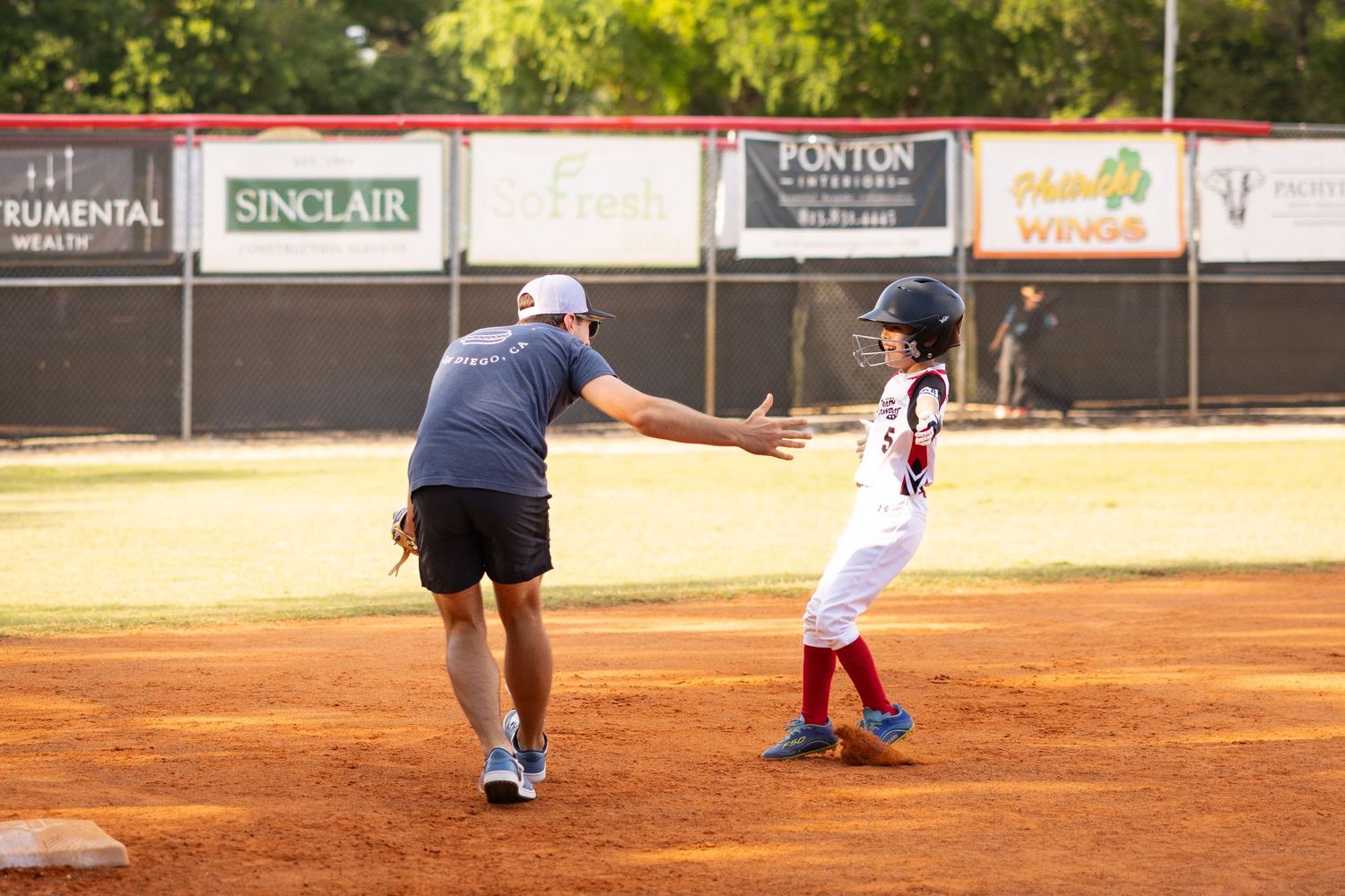 Coach high-fiving young baseball player on base during youth league game at outdoor diamond with sponsor banners.