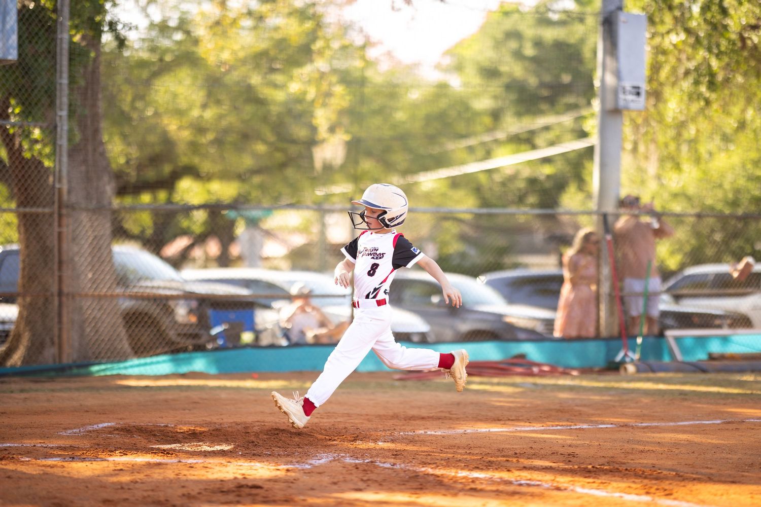 Young baseball player running bases in full uniform on a red dirt field during a sunny youth league game.