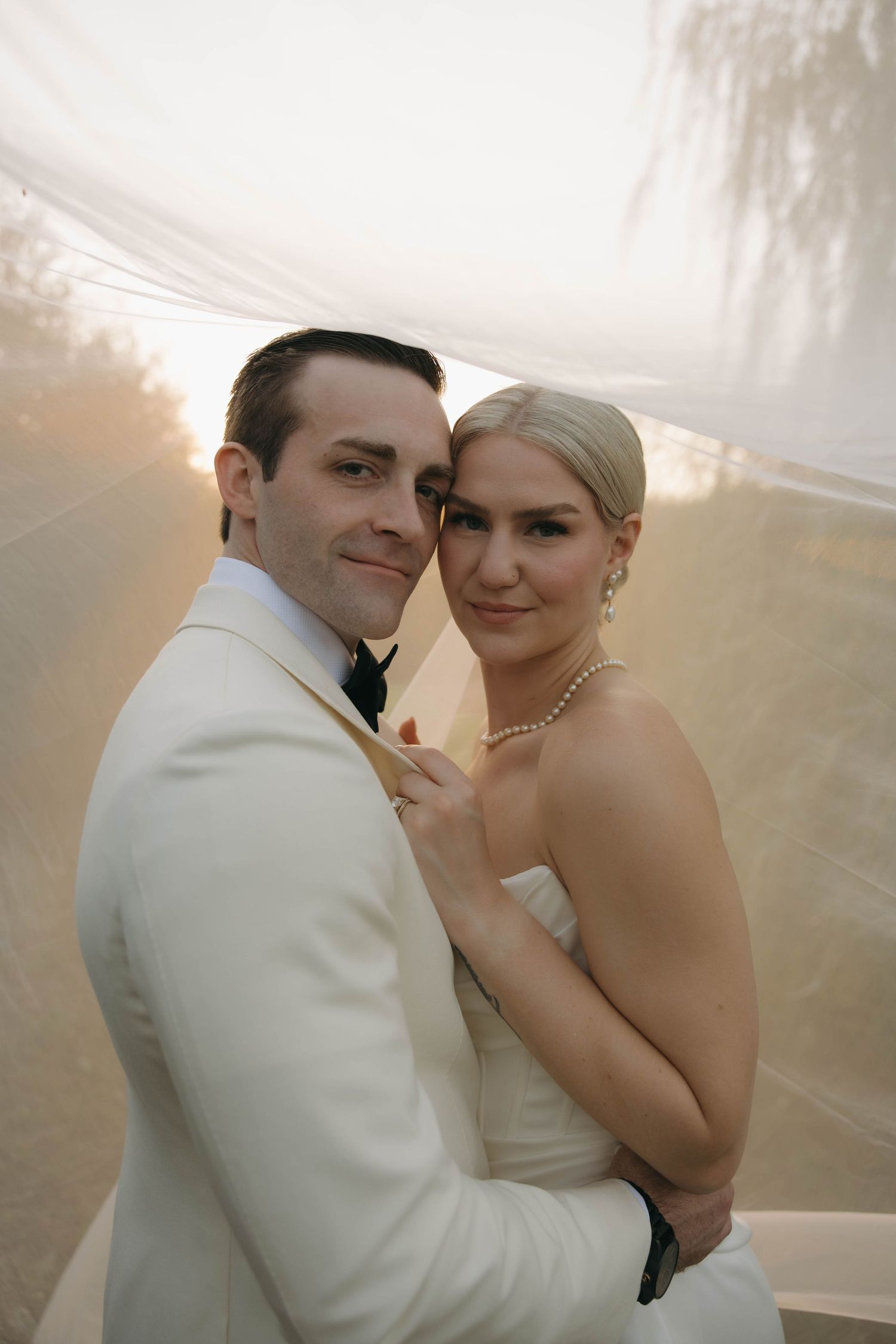A romantic wedding couple embraces in soft sunlight under a white veil during their special day.