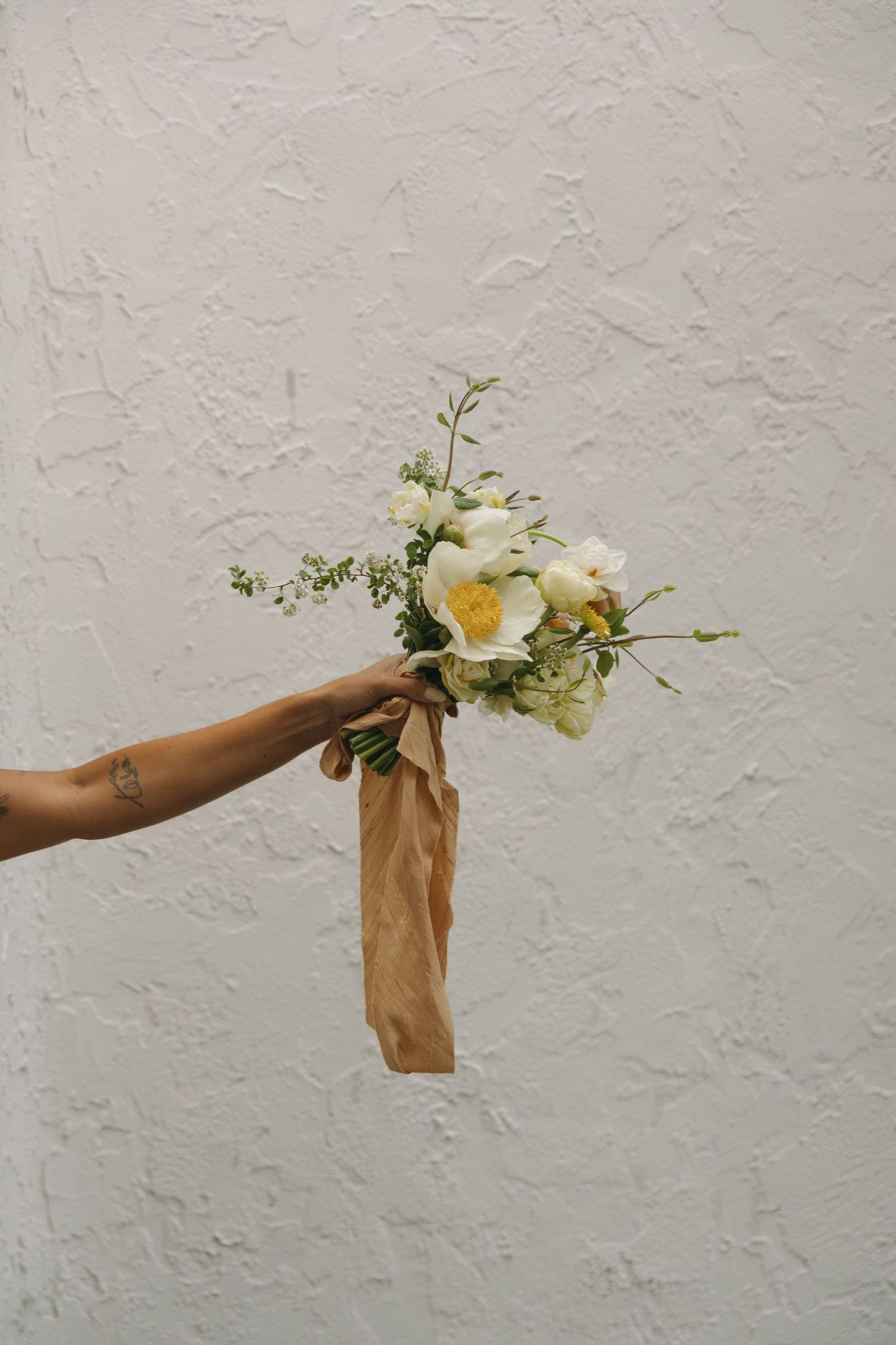 An outstretched arm holds a delicate bouquet of white flowers and greenery wrapped in brown kraft paper against a textured wall.