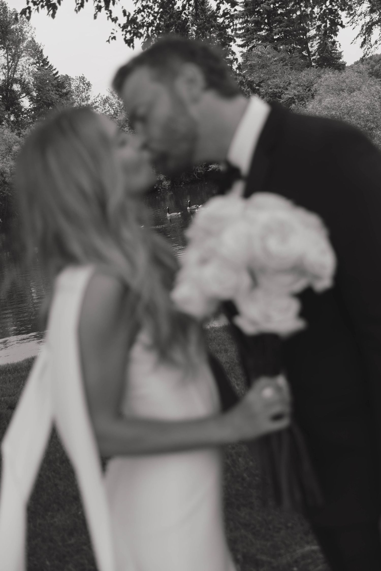 Black and white romantic wedding photo showing a couple kissing while holding a bouquet of light-colored flowers outdoors.