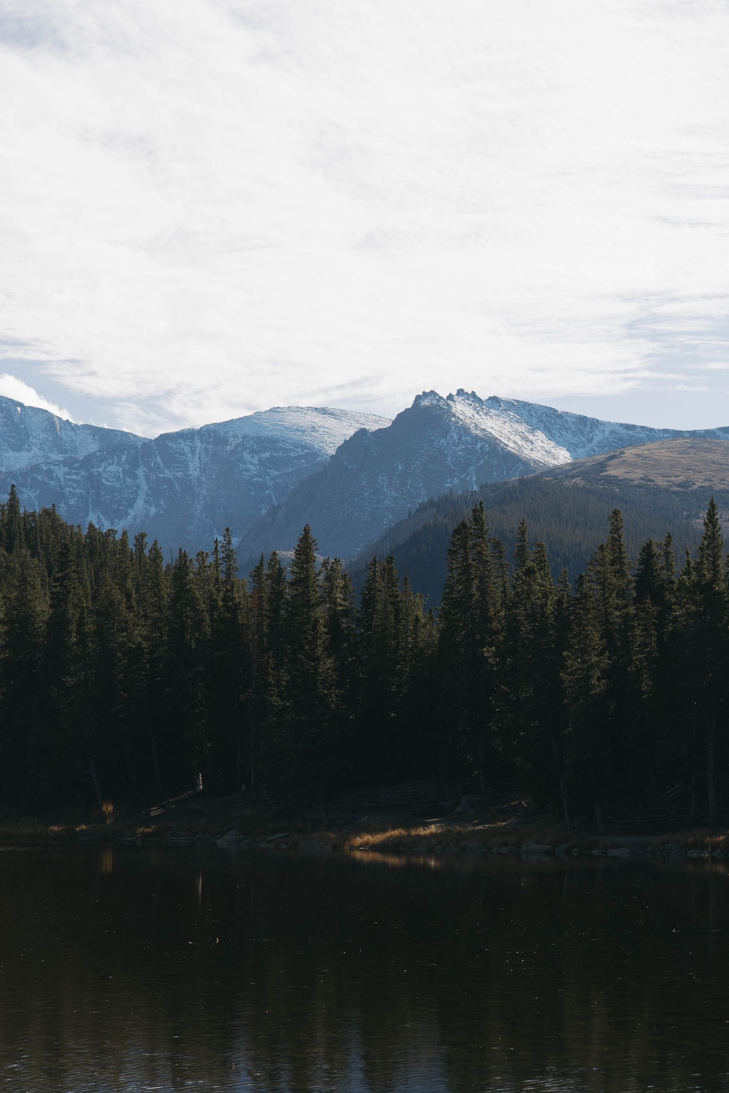 Snow-capped mountain peaks rise above a serene lake surrounded by dense evergreen forest in a wilderness landscape.