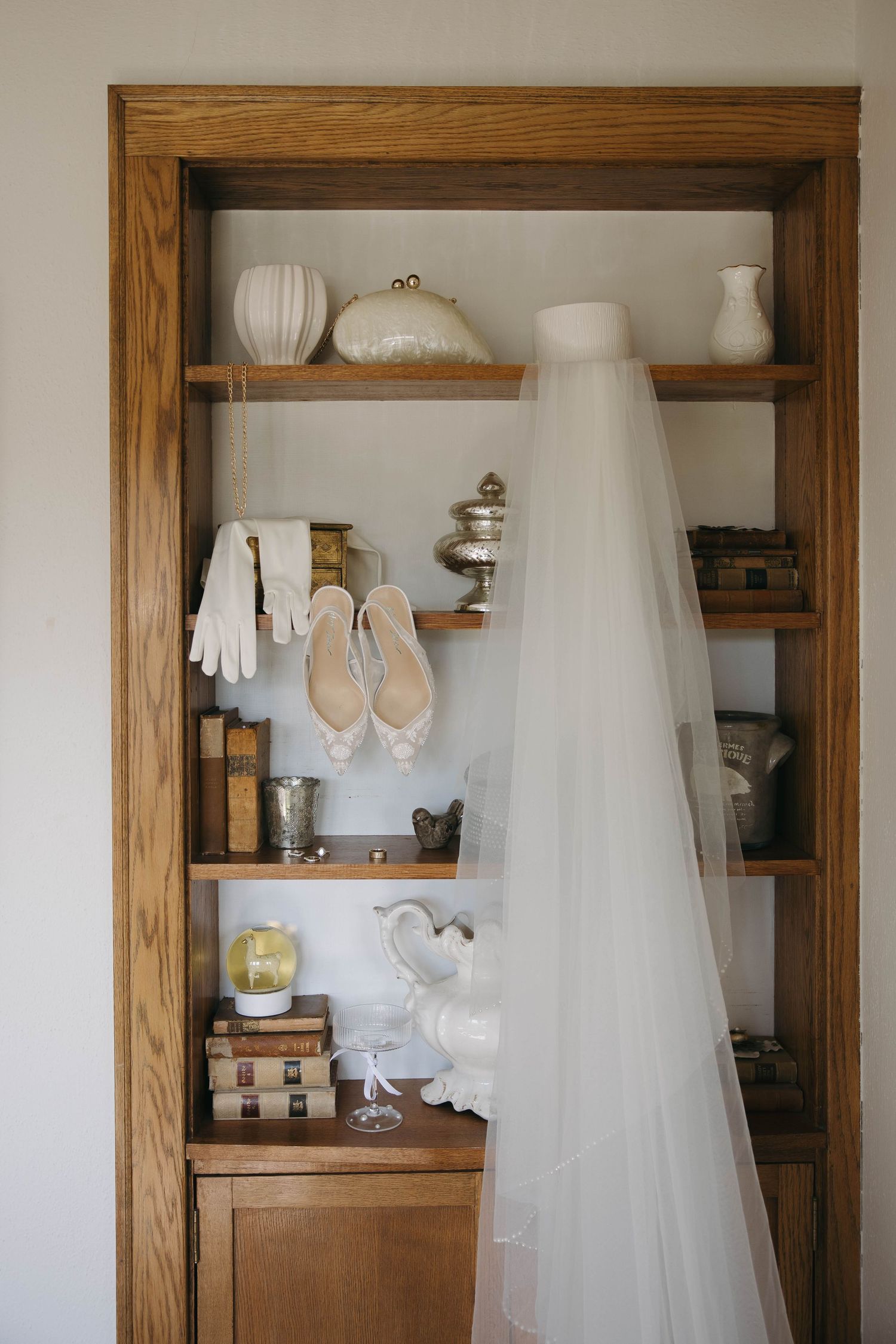 A wedding dress and accessories displayed in wooden built-in shelving with white ceramic decor pieces.