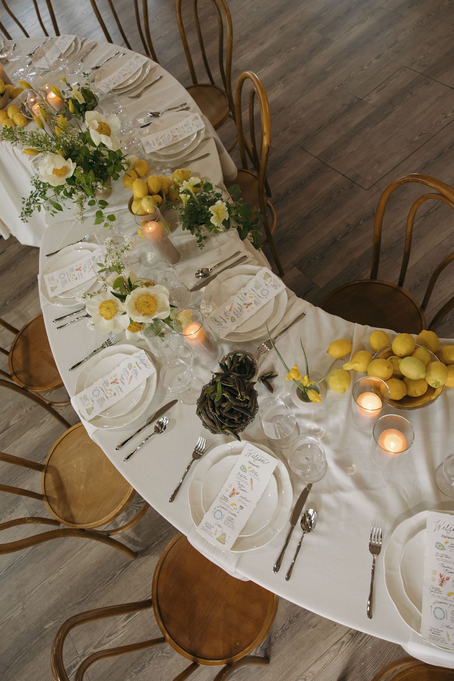 Elegant table setting with white linens, wooden chairs, lemon centerpieces and glowing candles on a curved dining table.