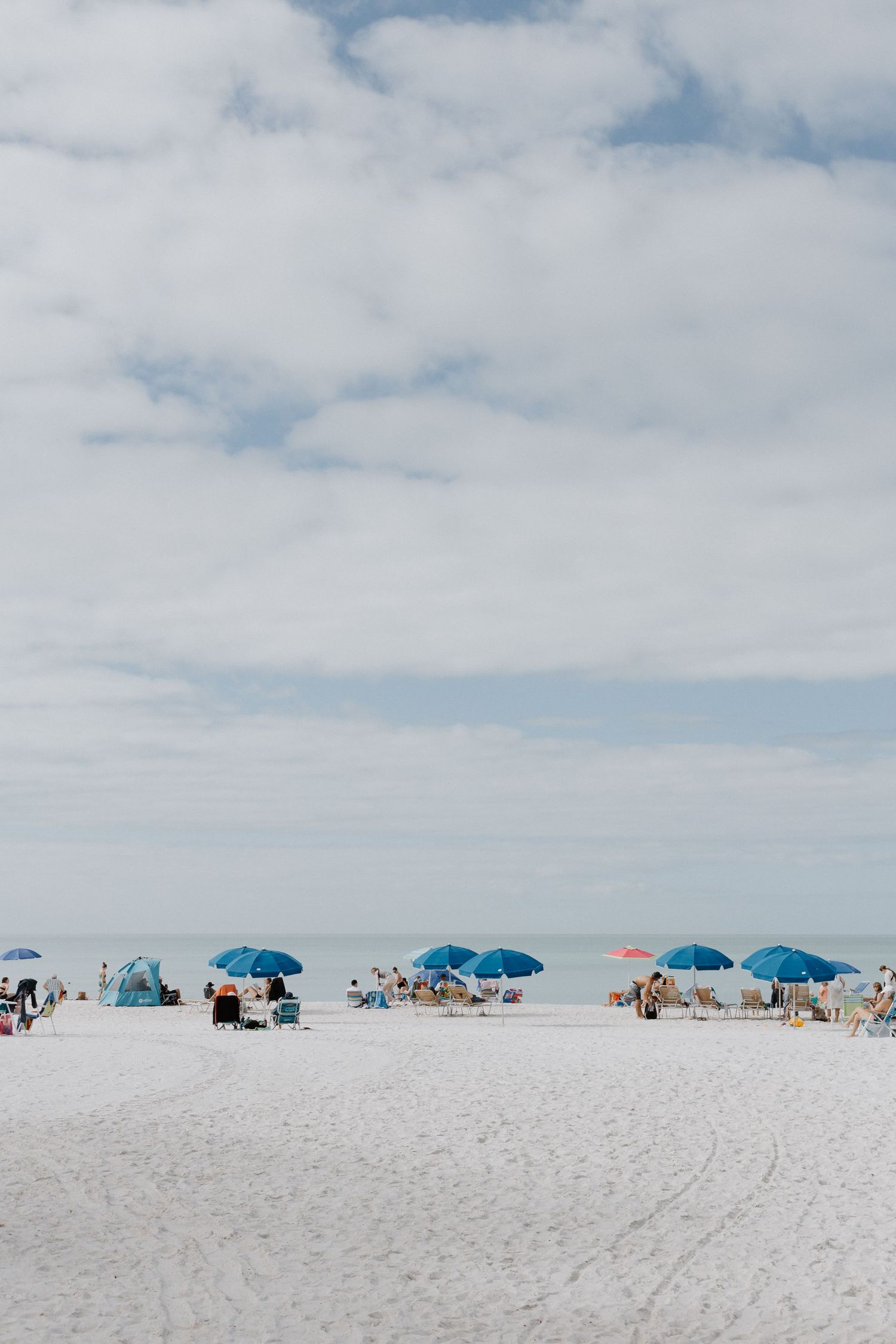 White sandy beach with scattered blue umbrellas and beachgoers under a cloudy summer sky.
