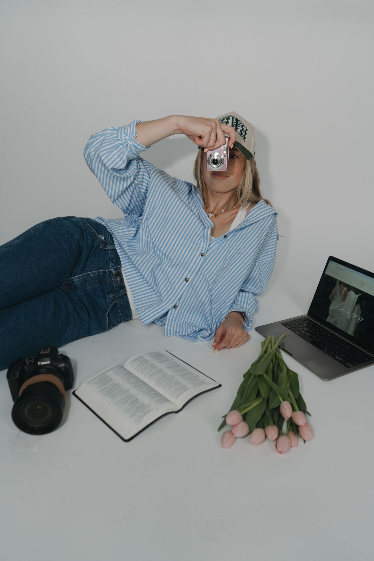 A person in a striped blue shirt lies on a white surface with a book, laptop, and green plant nearby.