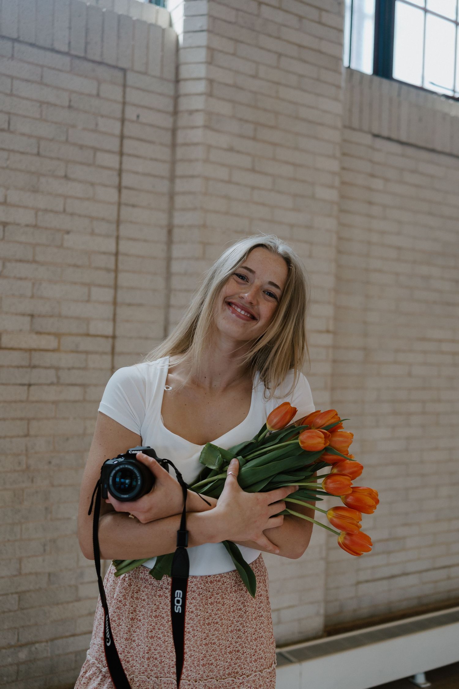 Colorado-based photographer in white shirt holding orange tulips and camera against brick wall.