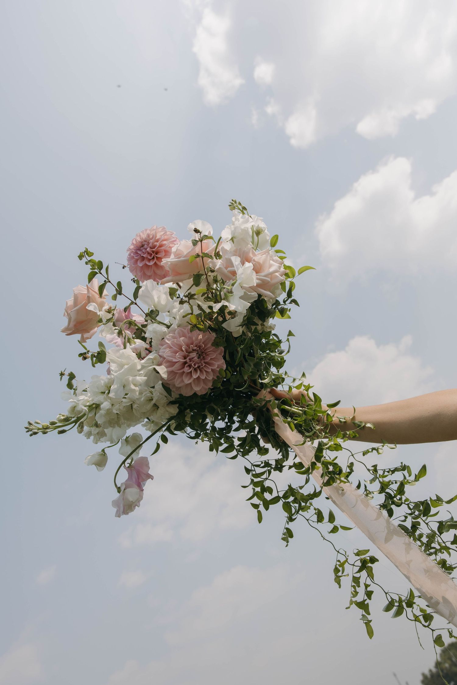 Hand holding a delicate floral arrangement with pink and white blooms against a cloudy sky background.