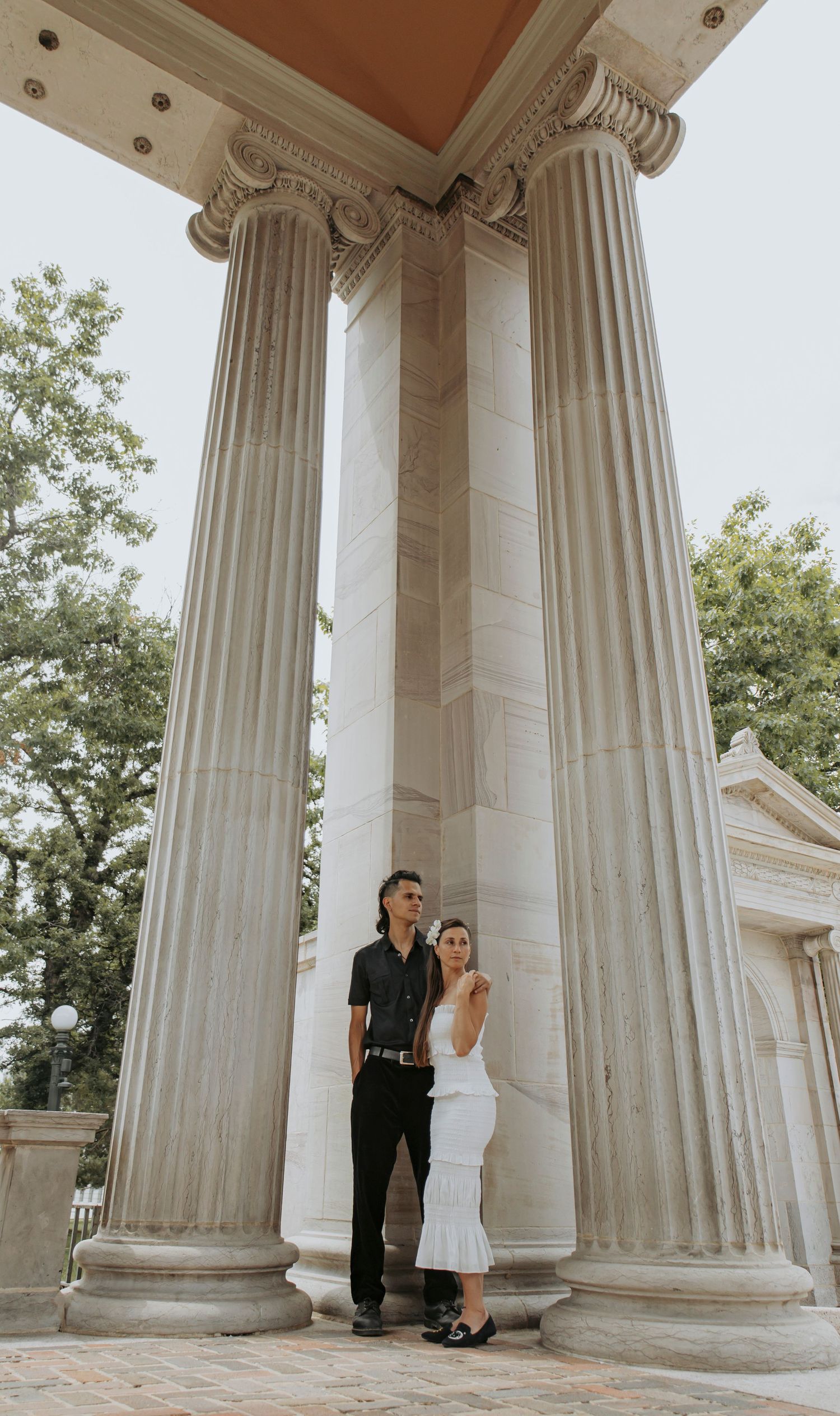 A couple in formal attire poses between tall classical Corinthian columns on a marble porch.