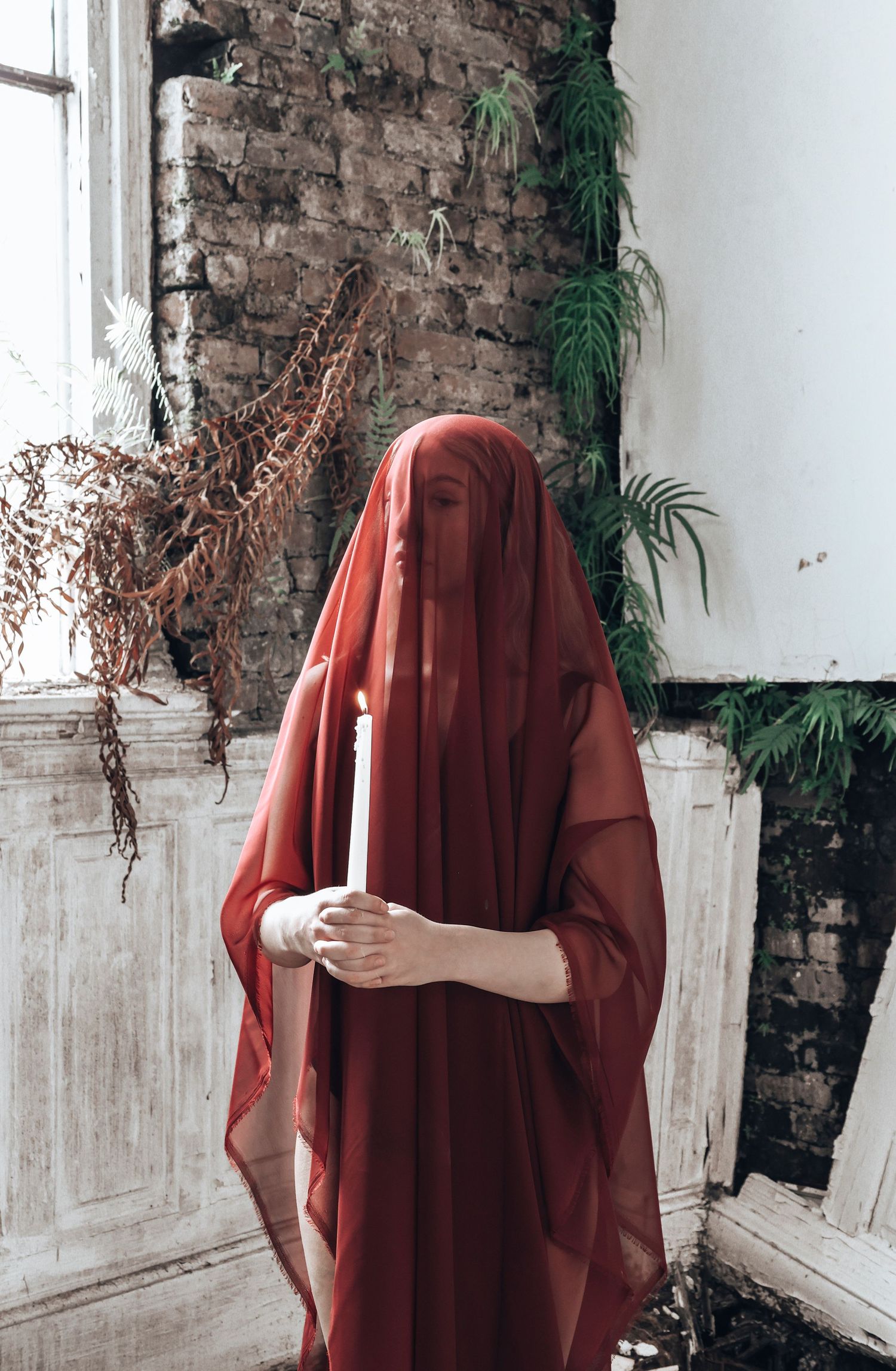A figure in a red cloak stands mysteriously against a rustic brick wall with dried flowers and hanging plants.