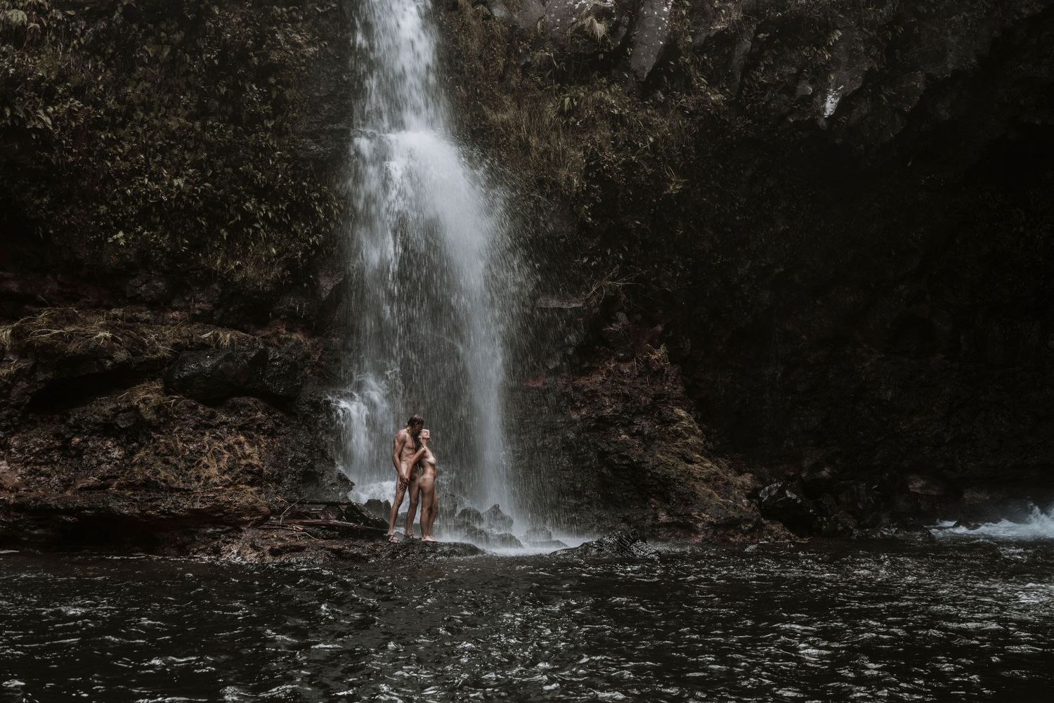 Two people stand in a dark rocky pool beneath a majestic cascading waterfall surrounded by cliffs.