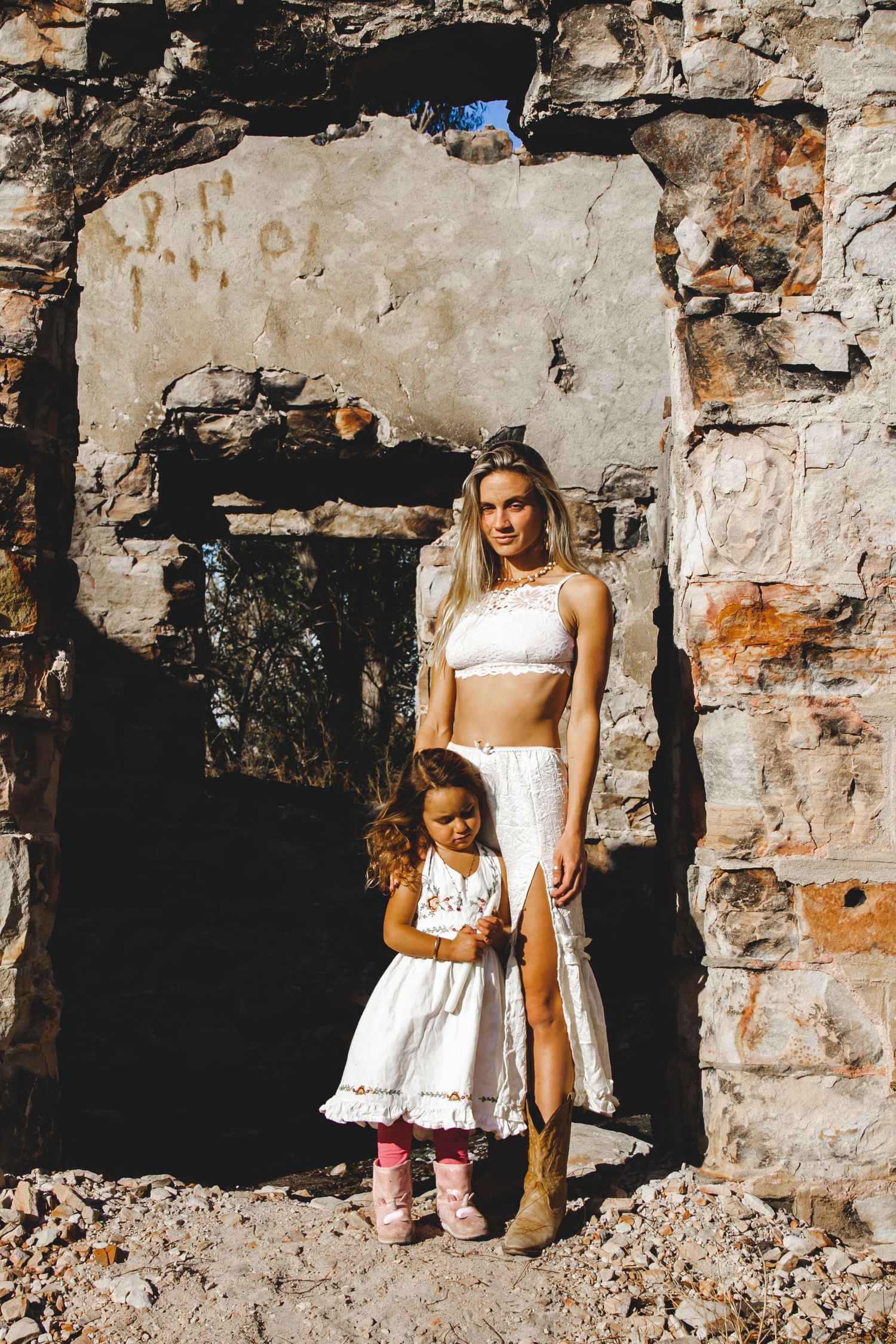 A mother and daughter in white outfits standing in a stone archway of an old building during golden hour lighting.