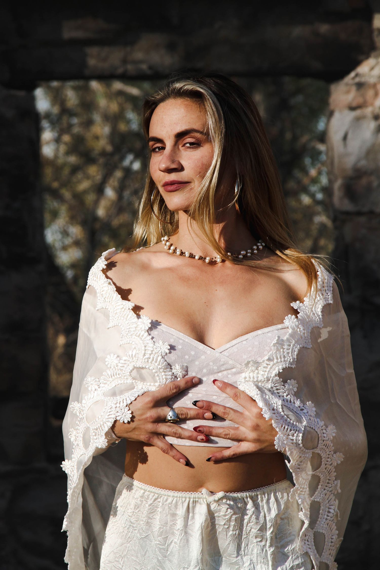 A feminine figure in a white lace crop top and pearl necklace poses against a shadowy stone background.