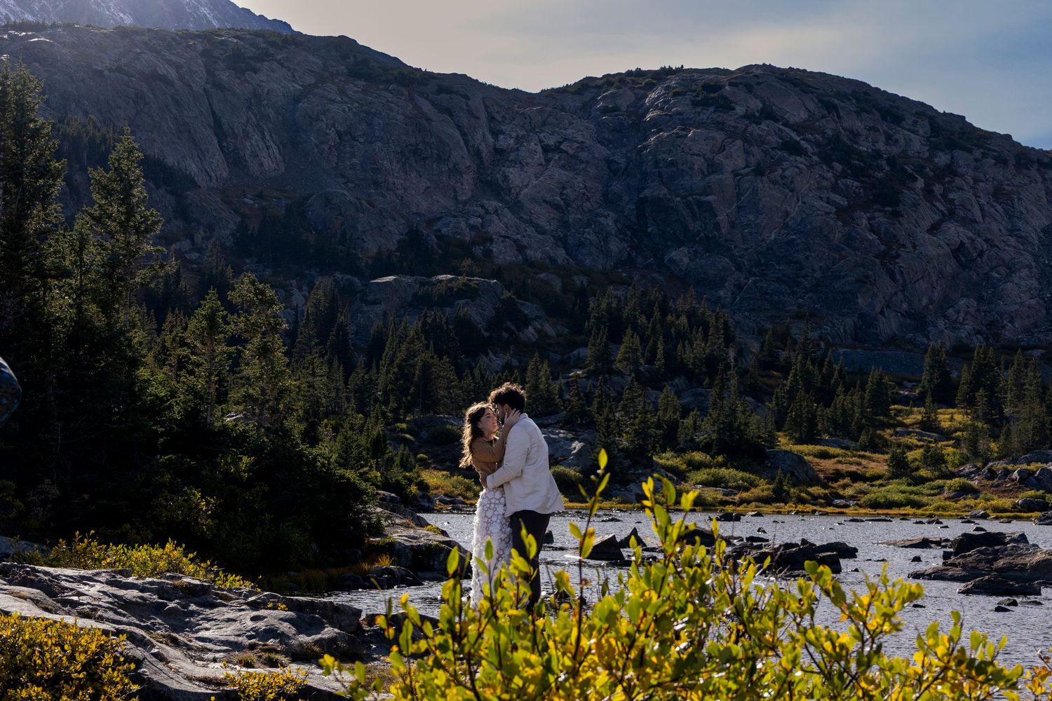 A couple embraces against a dramatic mountain landscape with evergreen trees and a flowing river on a sunny day.