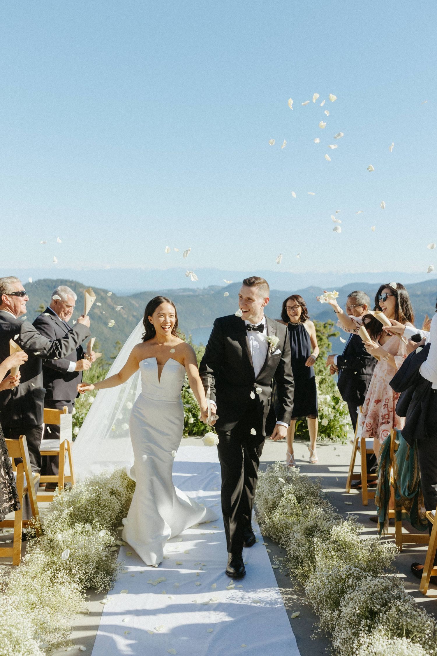 Newlyweds walk down outdoor aisle as guests toss white petals against mountain backdrop.
