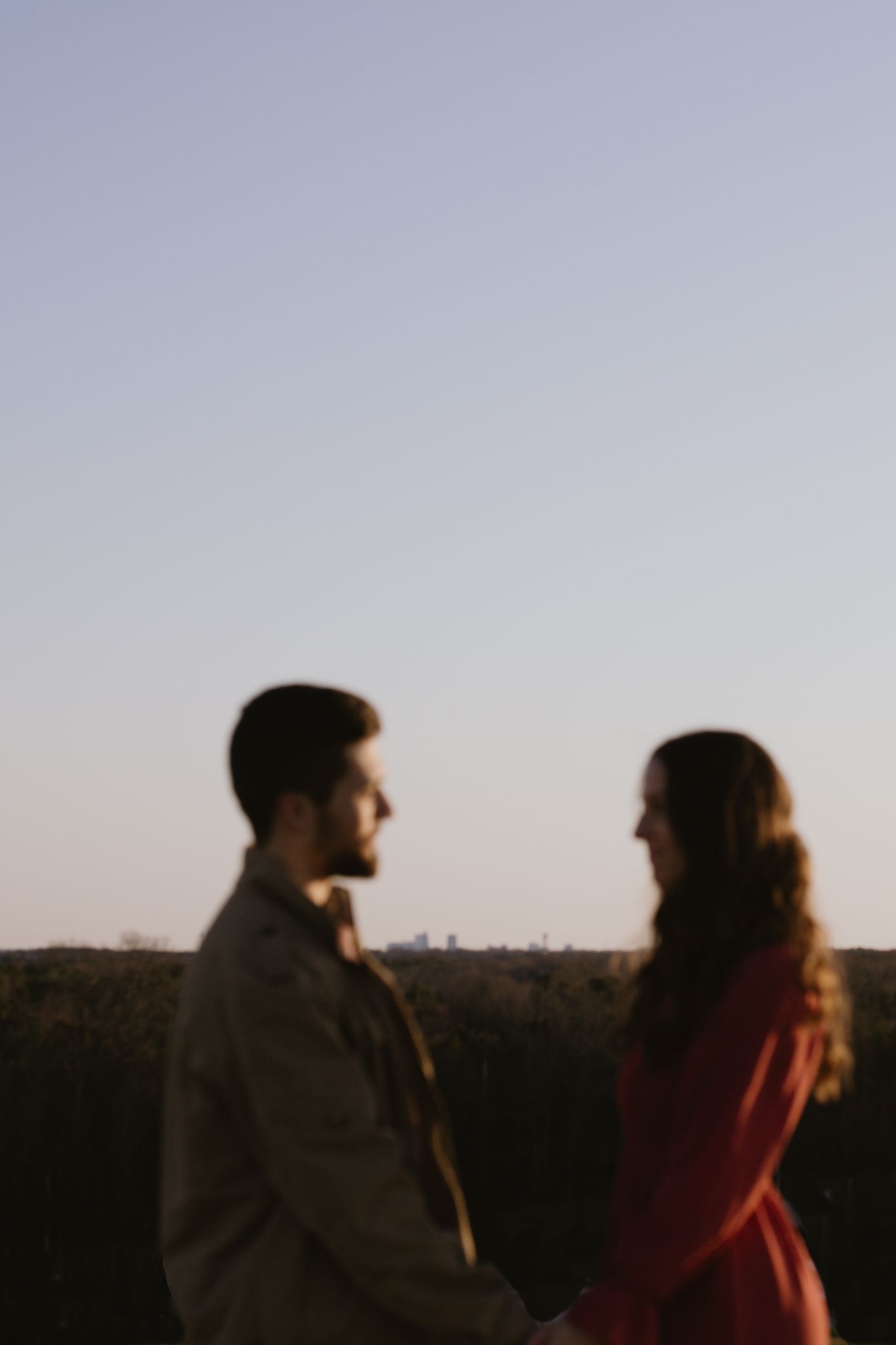 A couple is sharing an intimate glance holding hands. They are blurred out in front of the Raleigh skyline at a sunset p