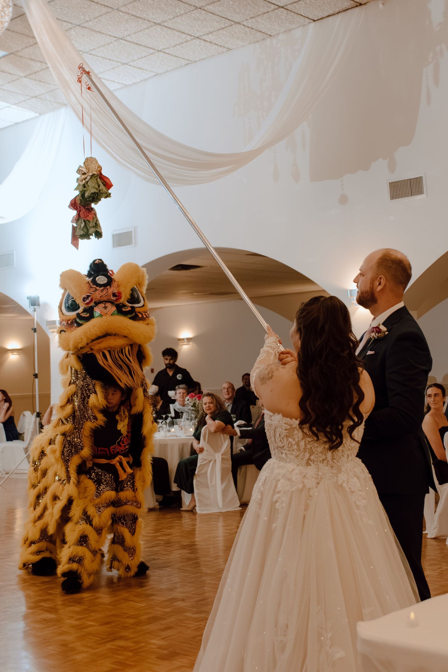 Bride and groom interact with a traditional Chinese lion dance performer at their elegant wedding reception.