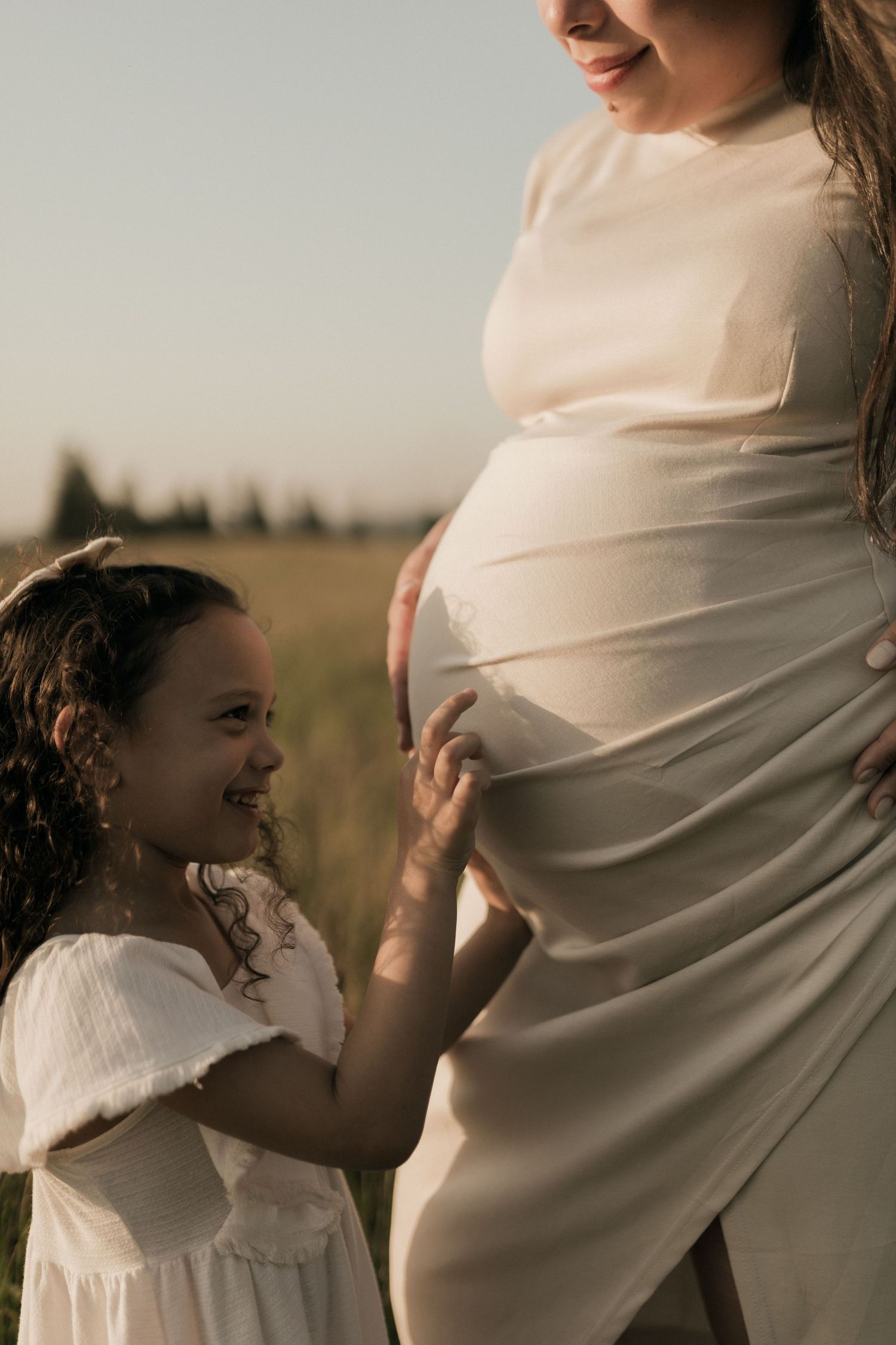 A young girl in a white dress smiles while touching a pregnant woman's belly outdoors in a sunny field.