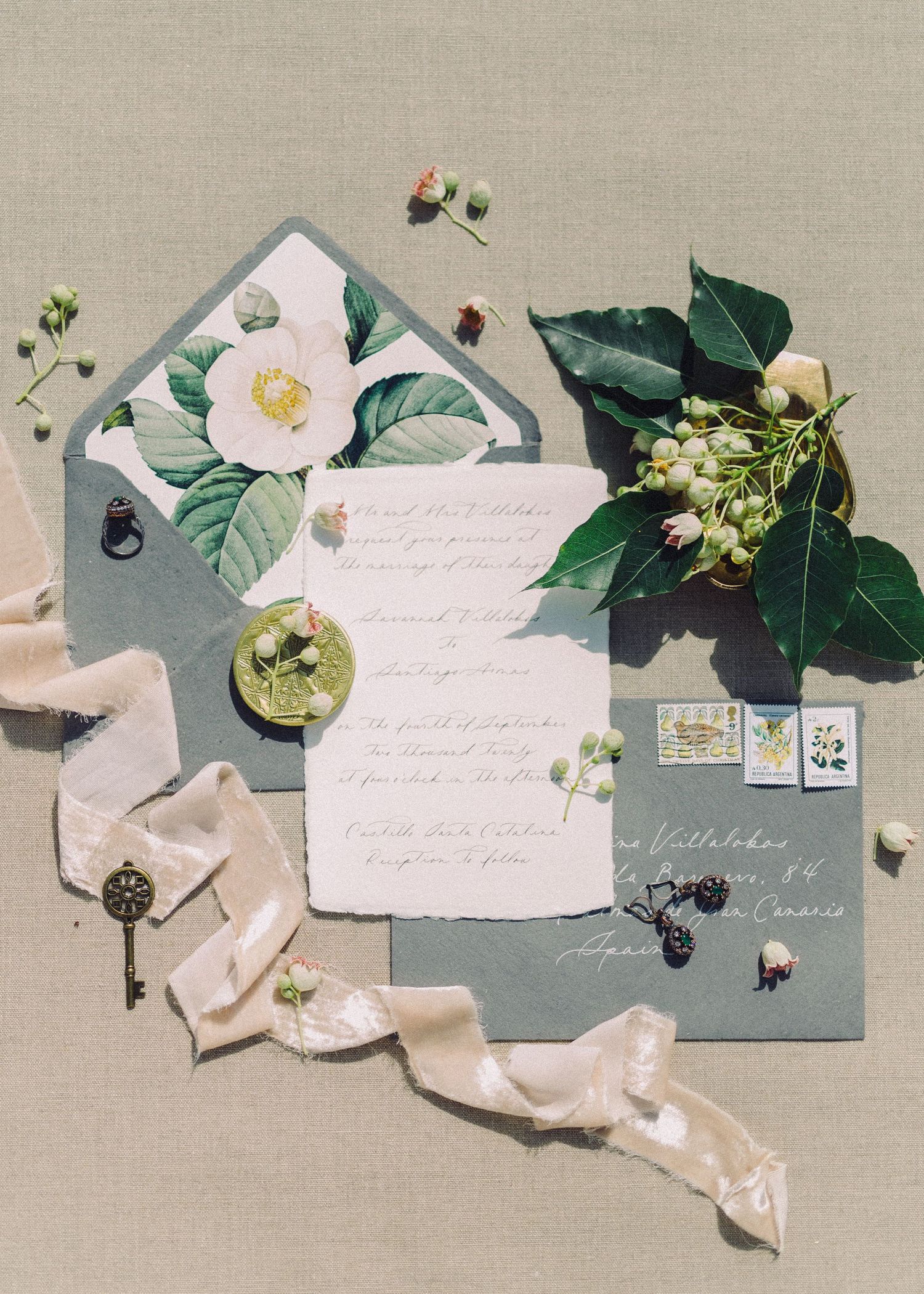 Elegant wedding invitation suite with dusty blue envelopes, floral liner, greenery, ribbon, and vintage key accents on linen.
