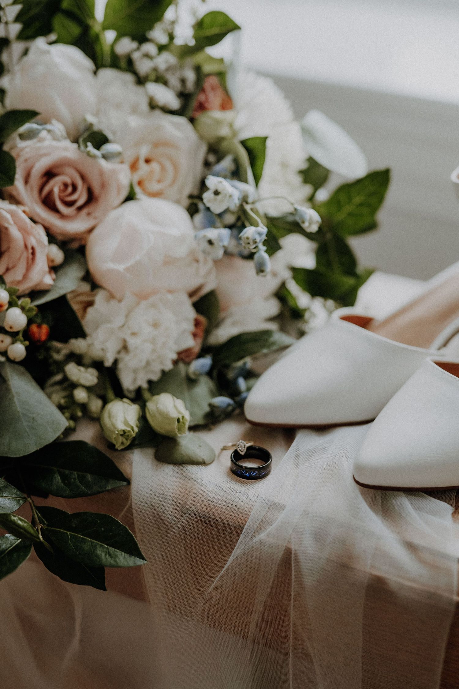 Elegant wedding flat lay with blush roses bouquet, white heels, and gold rings on soft tulle fabric.