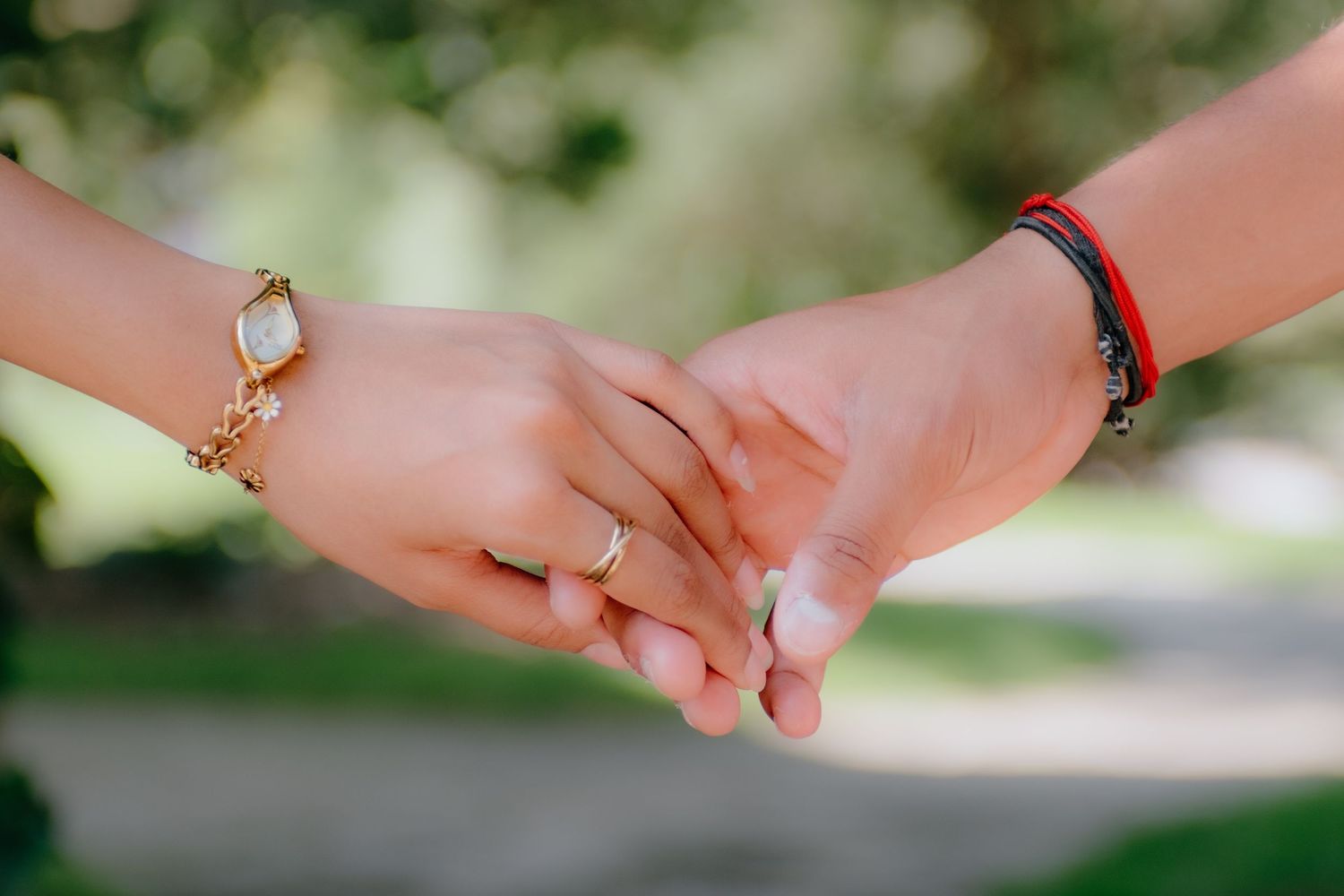 Two people holding hands gently, one wearing a gold bracelet and ring, the other a red string bracelet, with greenery in background.