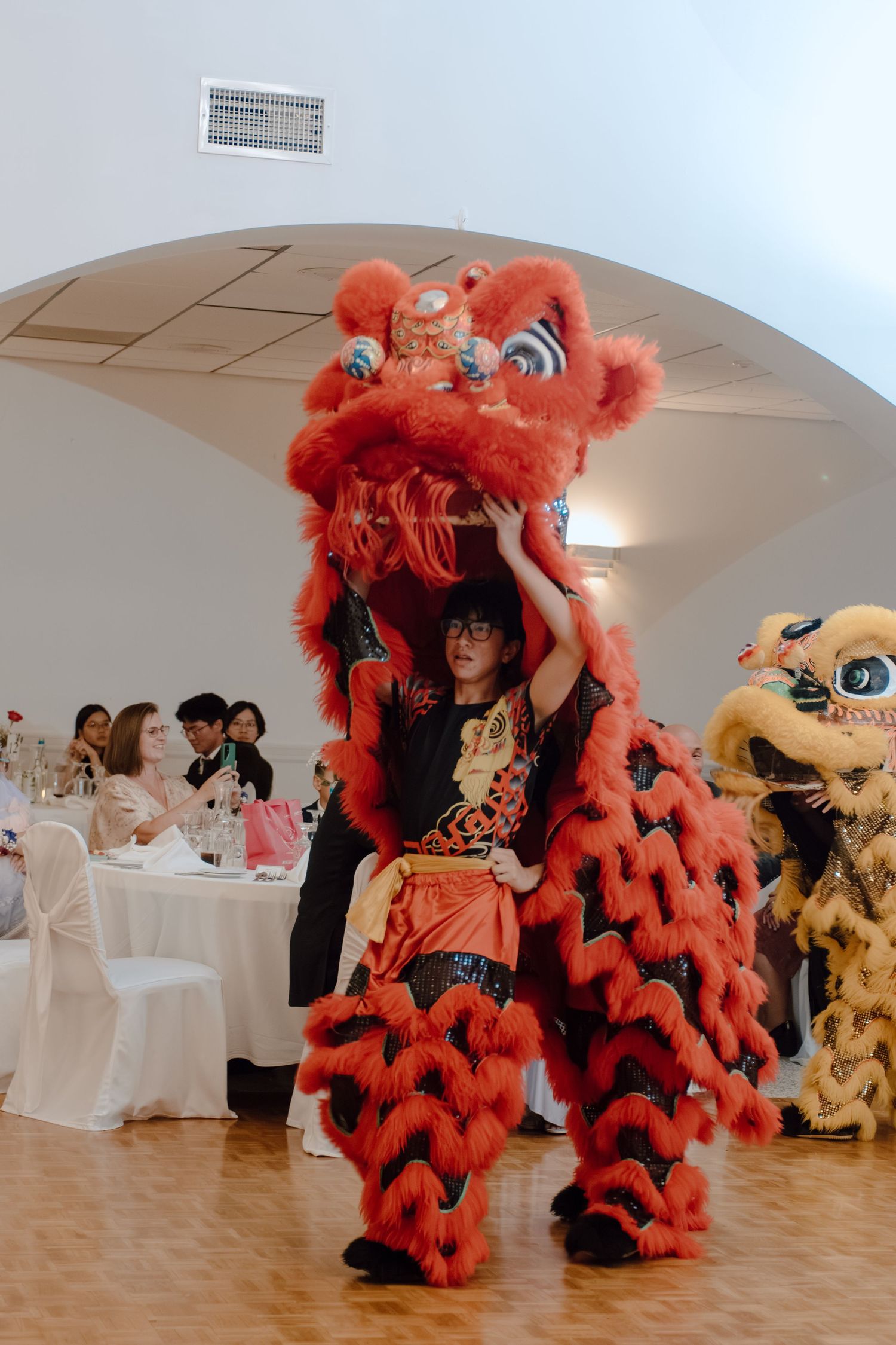 Performers in vibrant red and gold lion dance costumes entertain guests at a Chinese cultural celebration indoors.