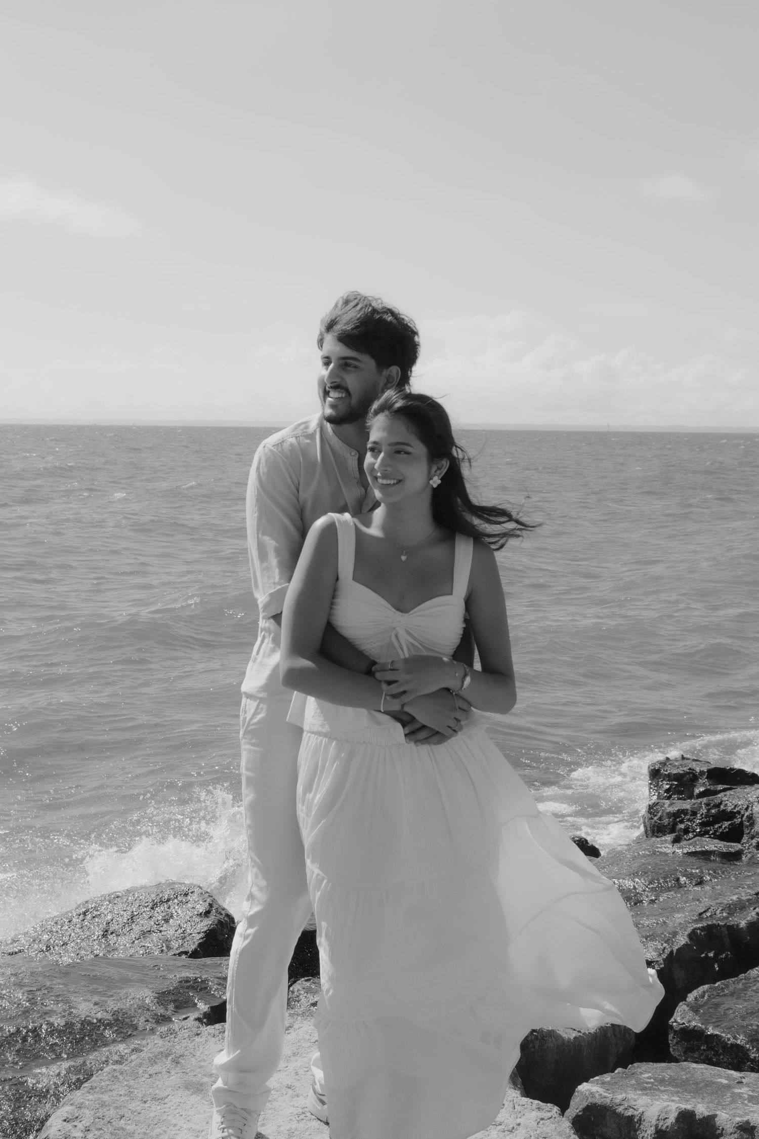 Black and white photo of a couple embracing on rocky ocean shore, woman in white dress, man in light shirt, waves behind them.