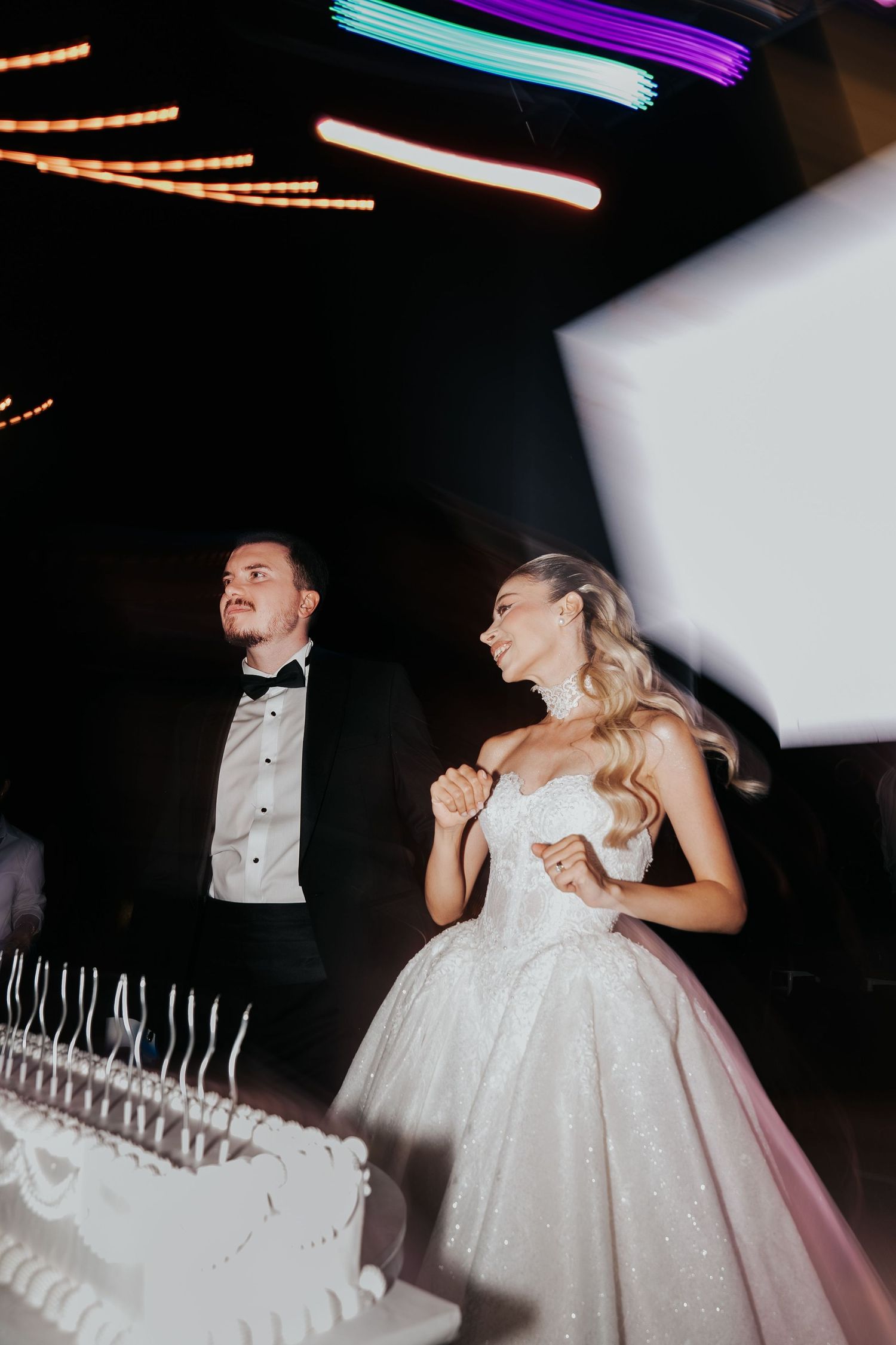 Bride in sparkly ball gown and groom in tuxedo stand beside wedding cake at elegant evening reception.