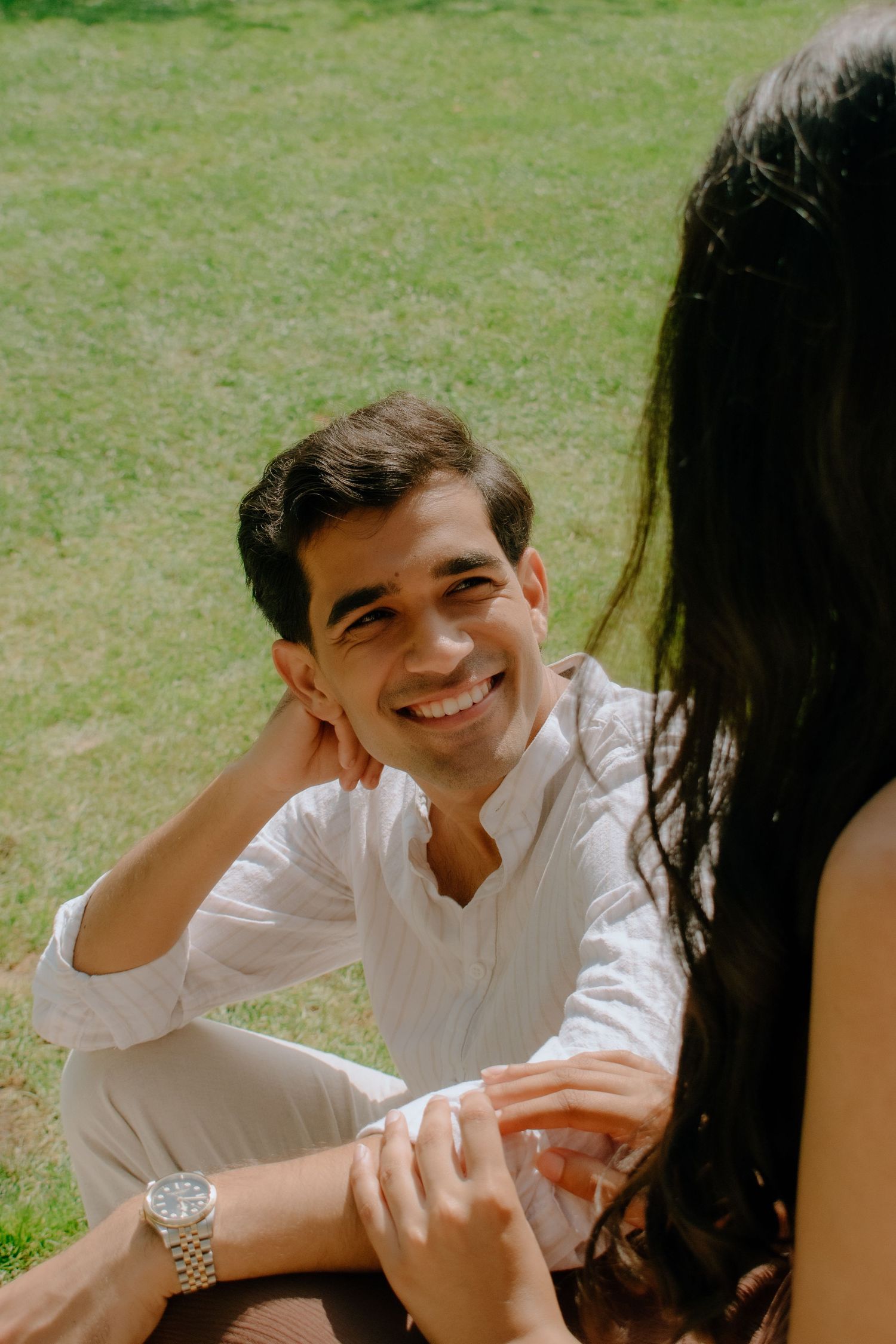 Smiling man in white shirt sitting on grass, holding hands with woman, enjoying a sunny outdoor date.