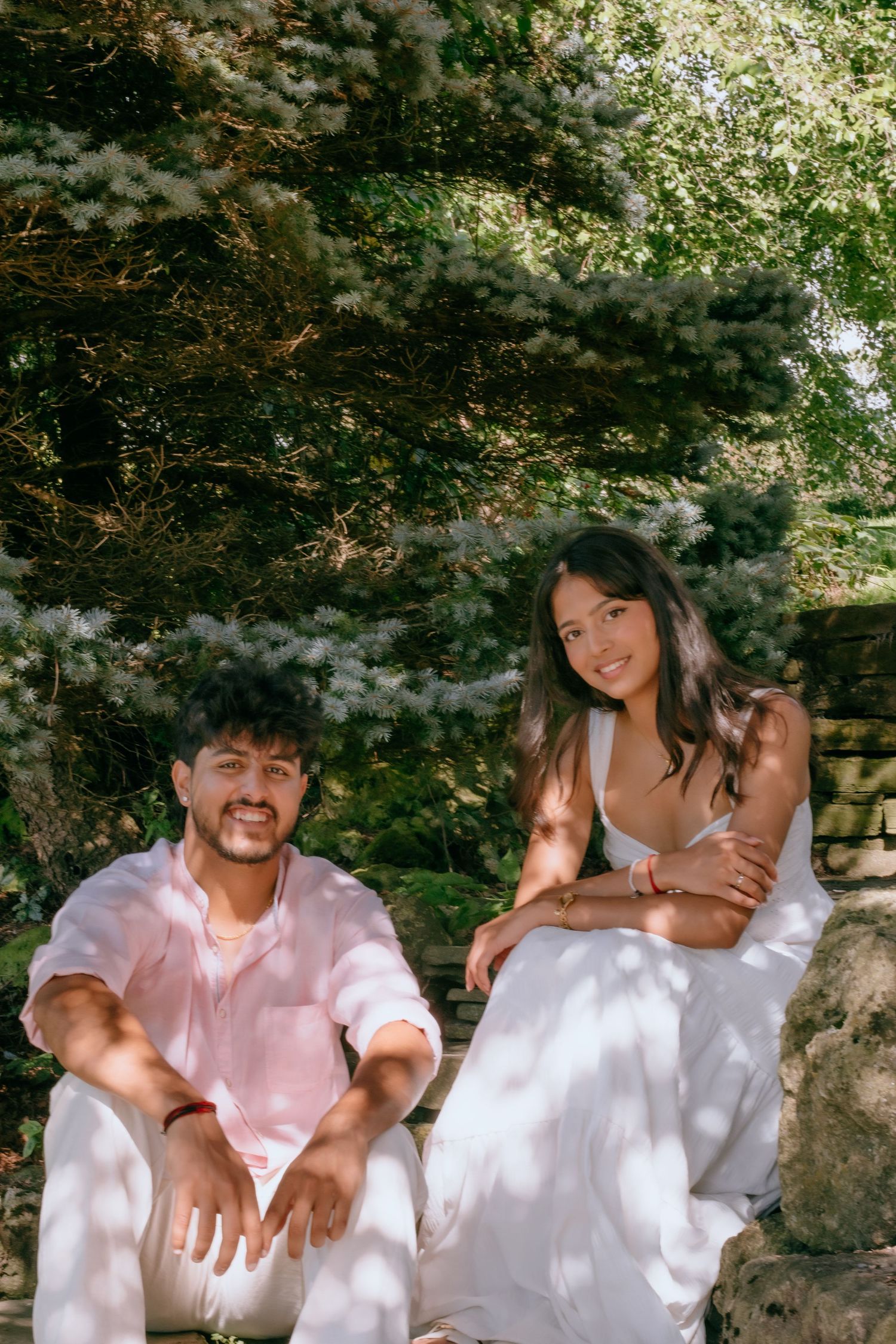 A young couple sits outdoors in a lush garden, smiling, wearing white and pink summer outfits.