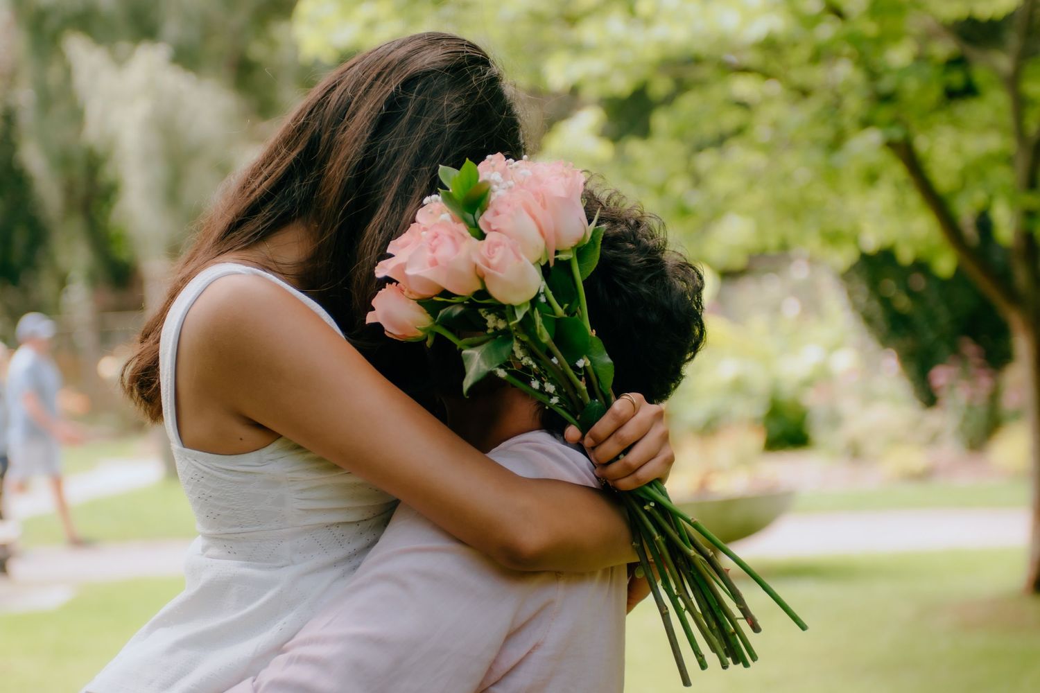 Two people embracing outdoors, one holding a bouquet of pink roses in a lush green garden setting.