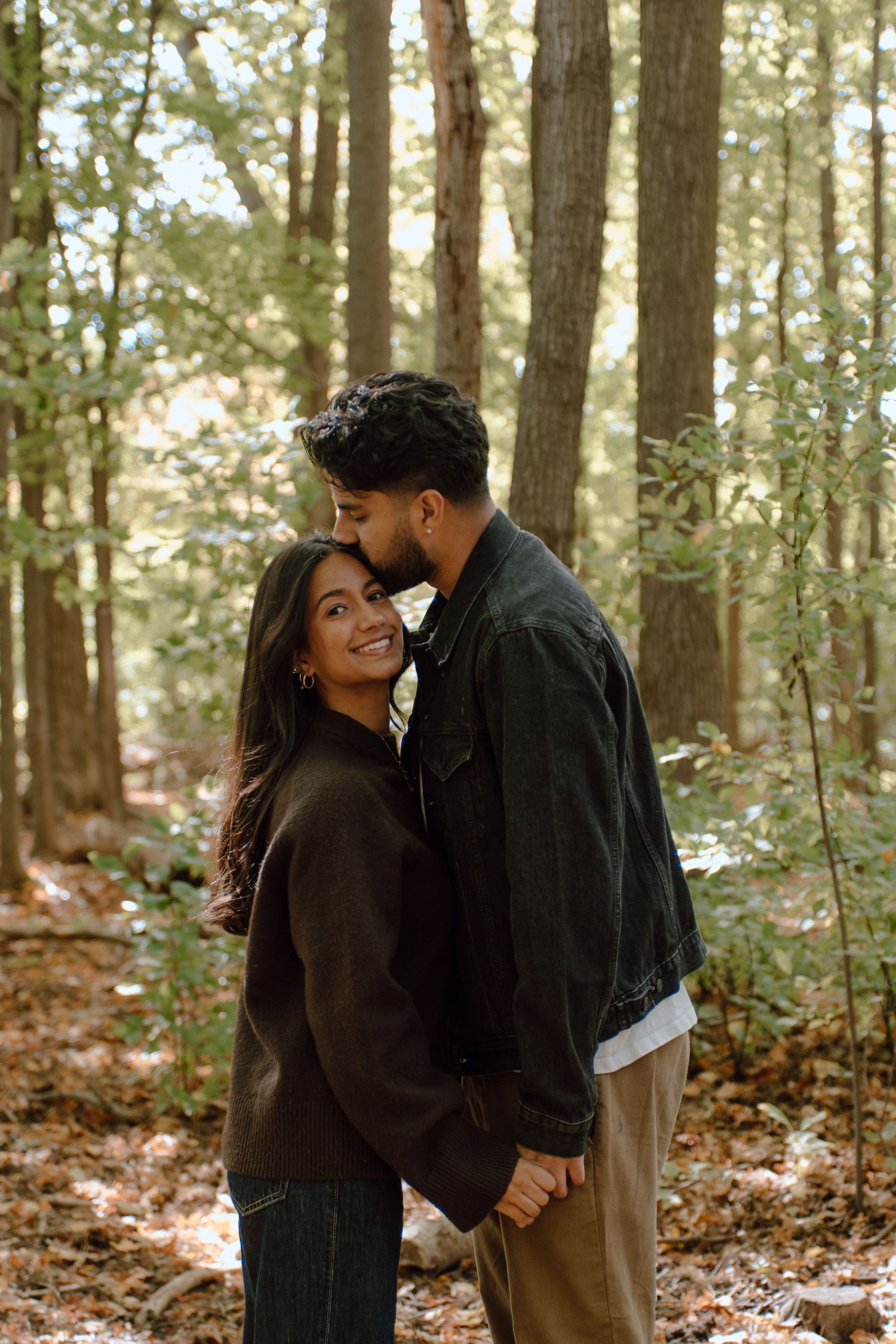 Couple holding hands and sharing a romantic moment in a sunlit forest, woman smiling at camera.