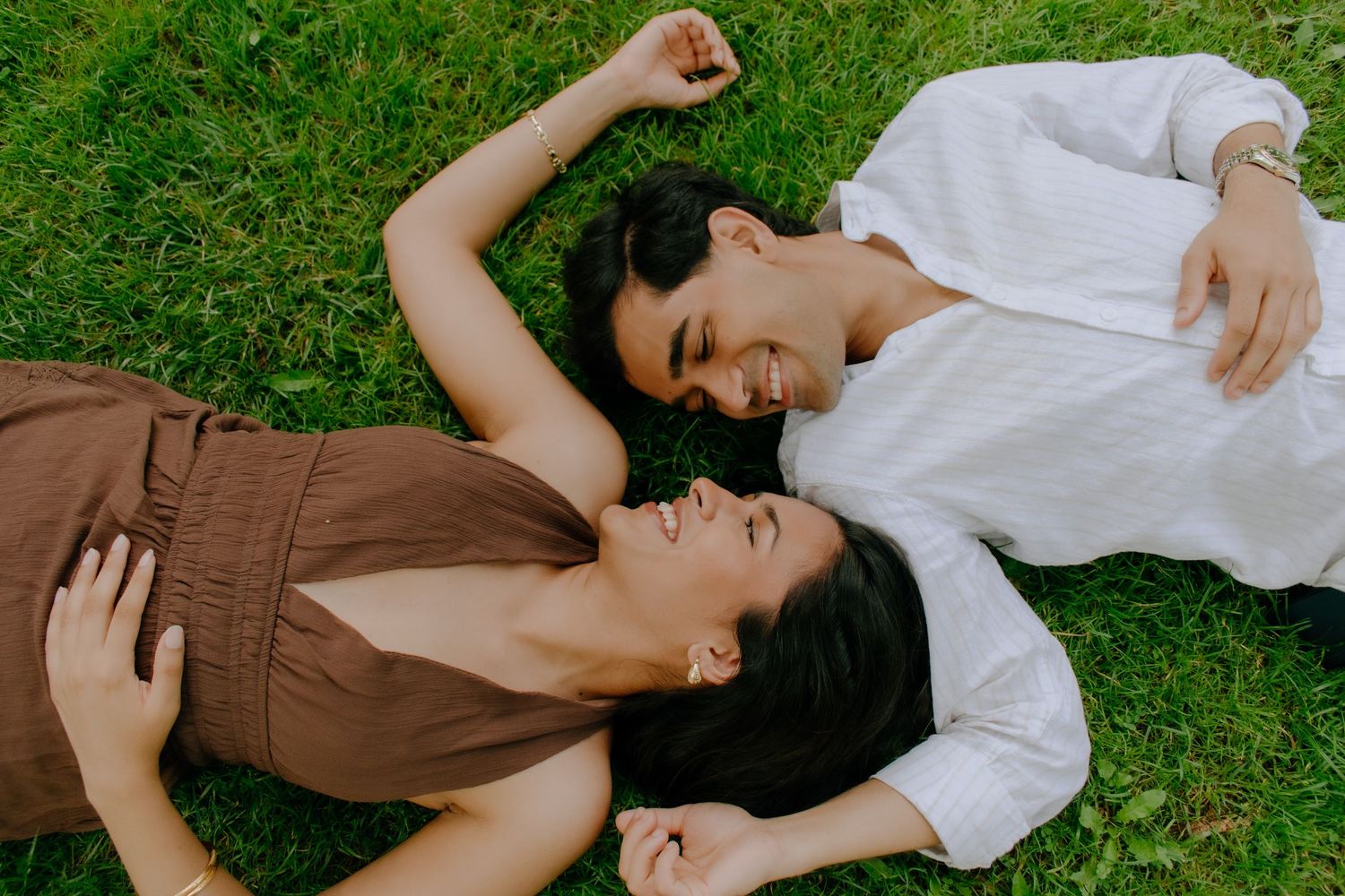 Two young women laughing and lying on green grass together, enjoying a sunny outdoor moment.
