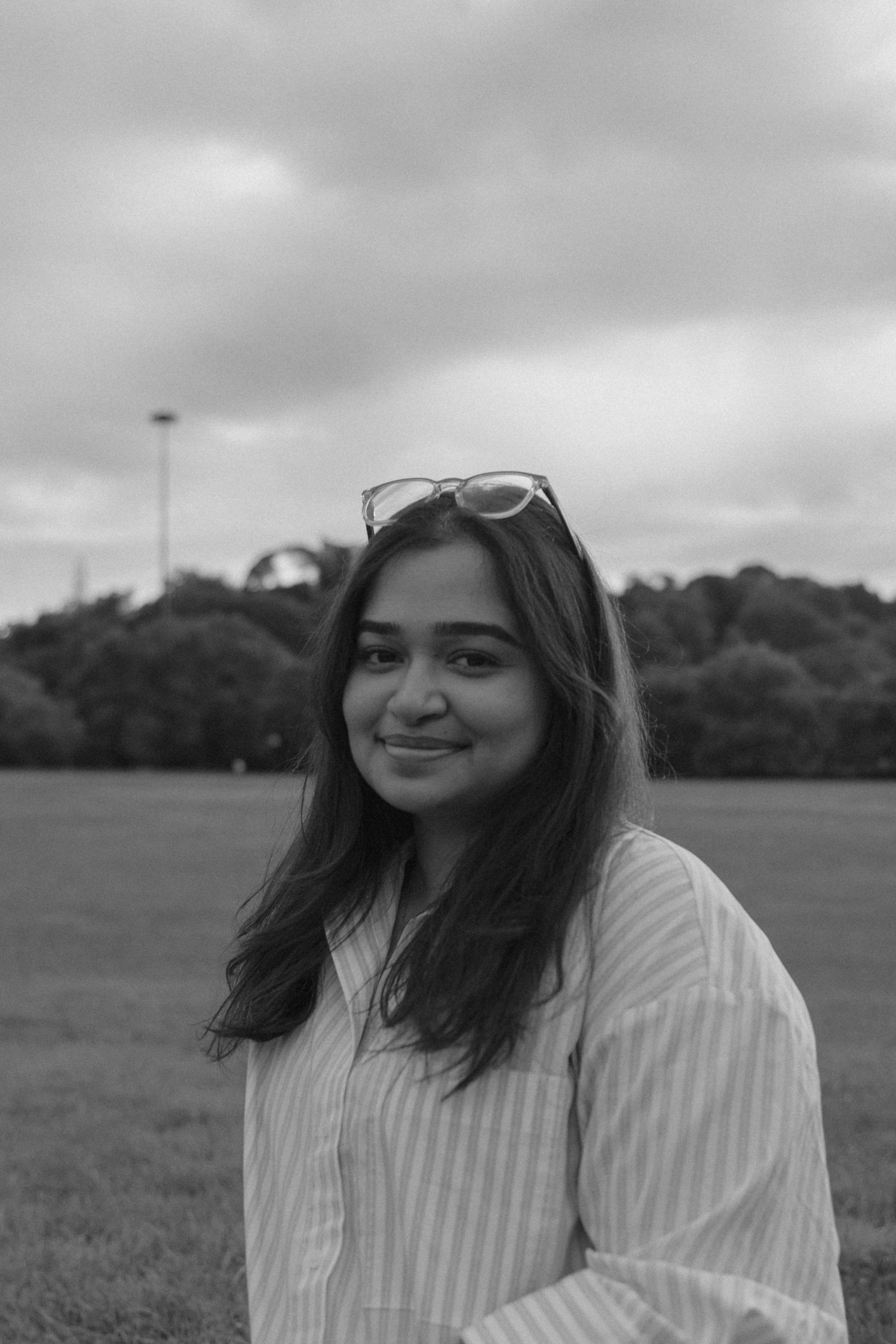 Black and white portrait of a young woman with sunglasses on head, smiling outdoors in a park.