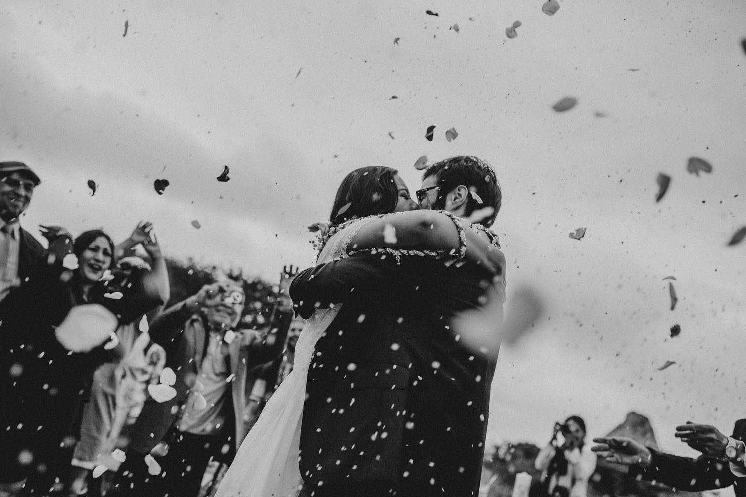 Black and white photo of newlyweds embracing amid confetti celebration outdoors.
