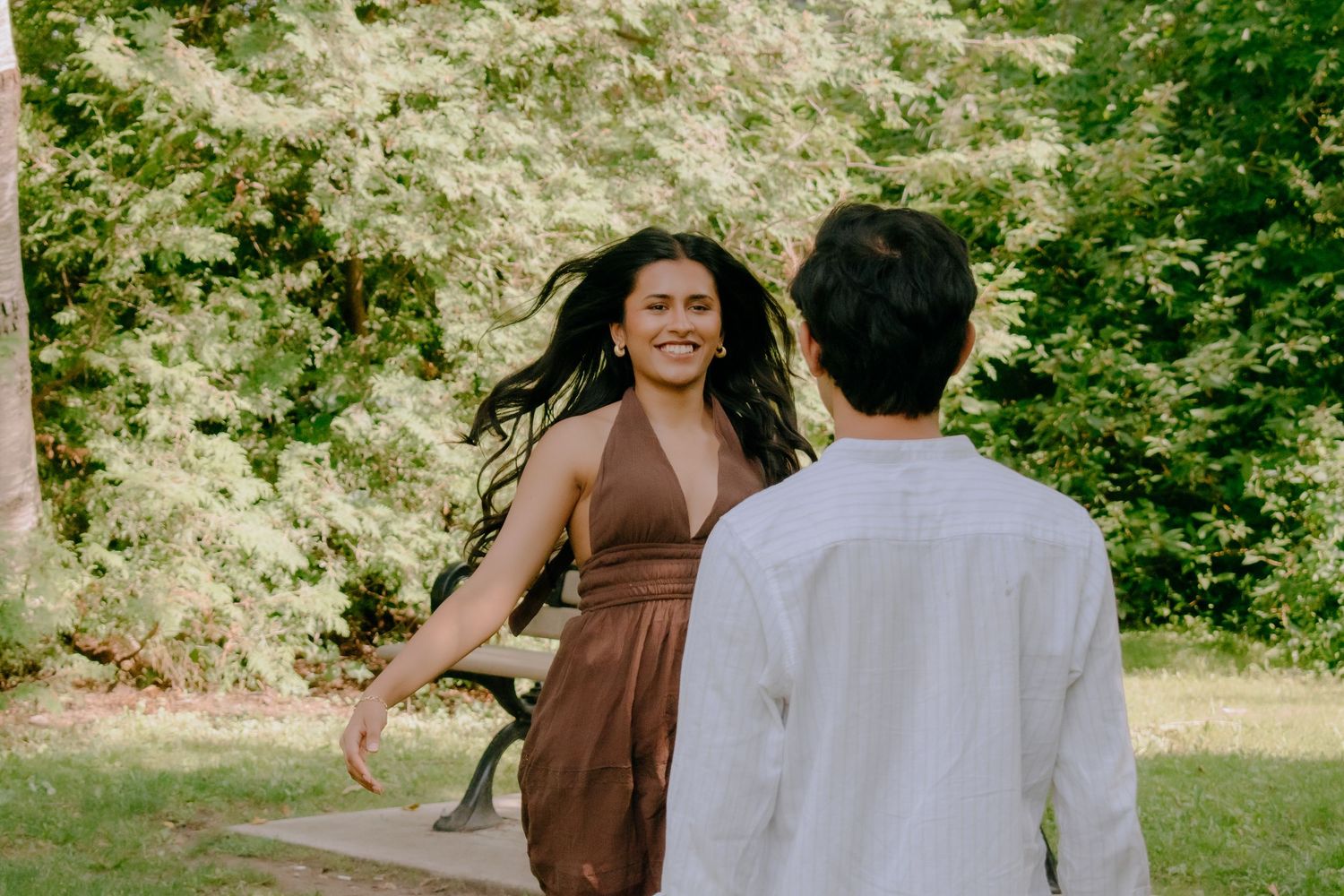 A smiling woman in a brown dress twirls in a park while a man in a white shirt watches her.