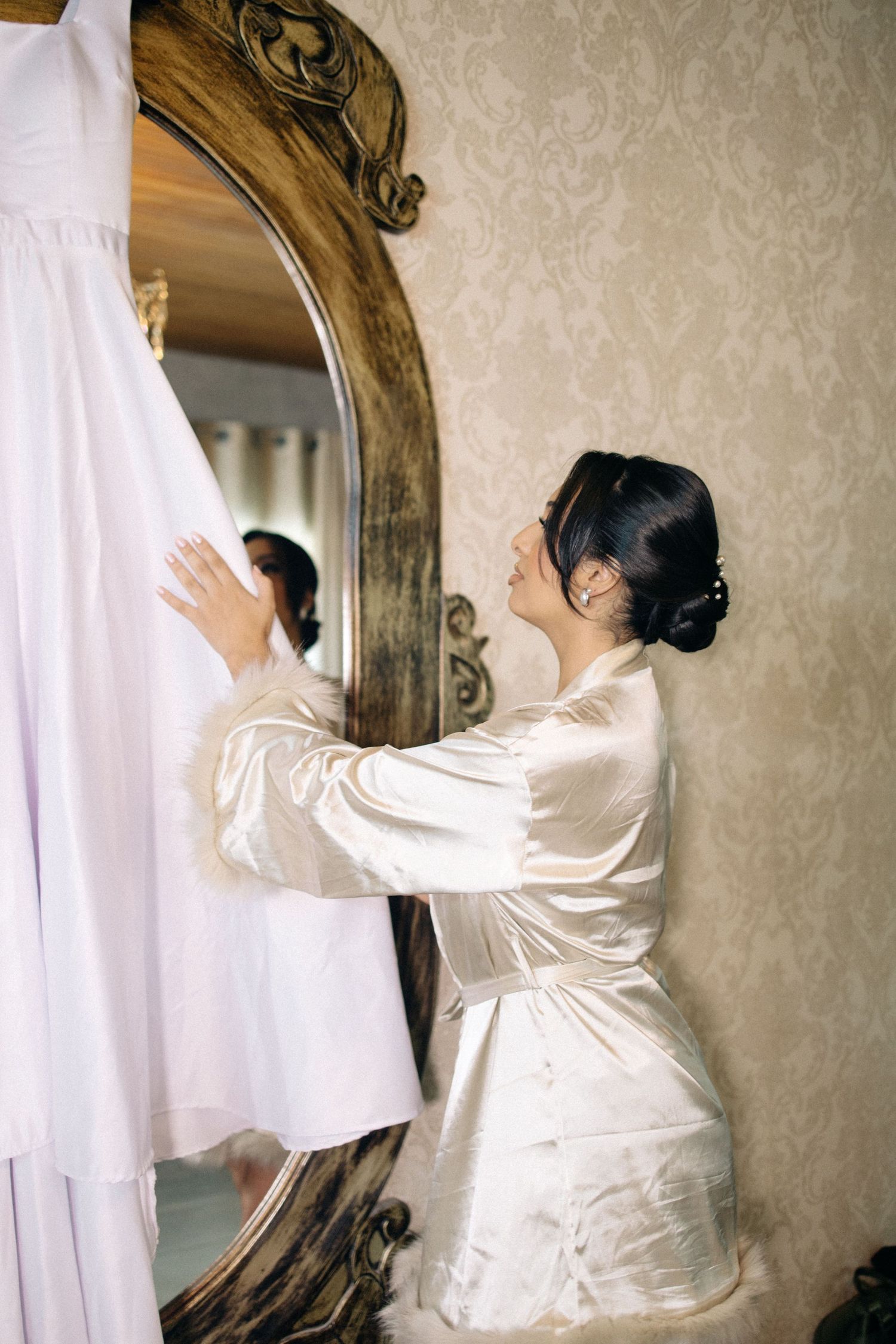 Bride in satin robe kneeling before ornate mirror, touching her white wedding dress hanging nearby.