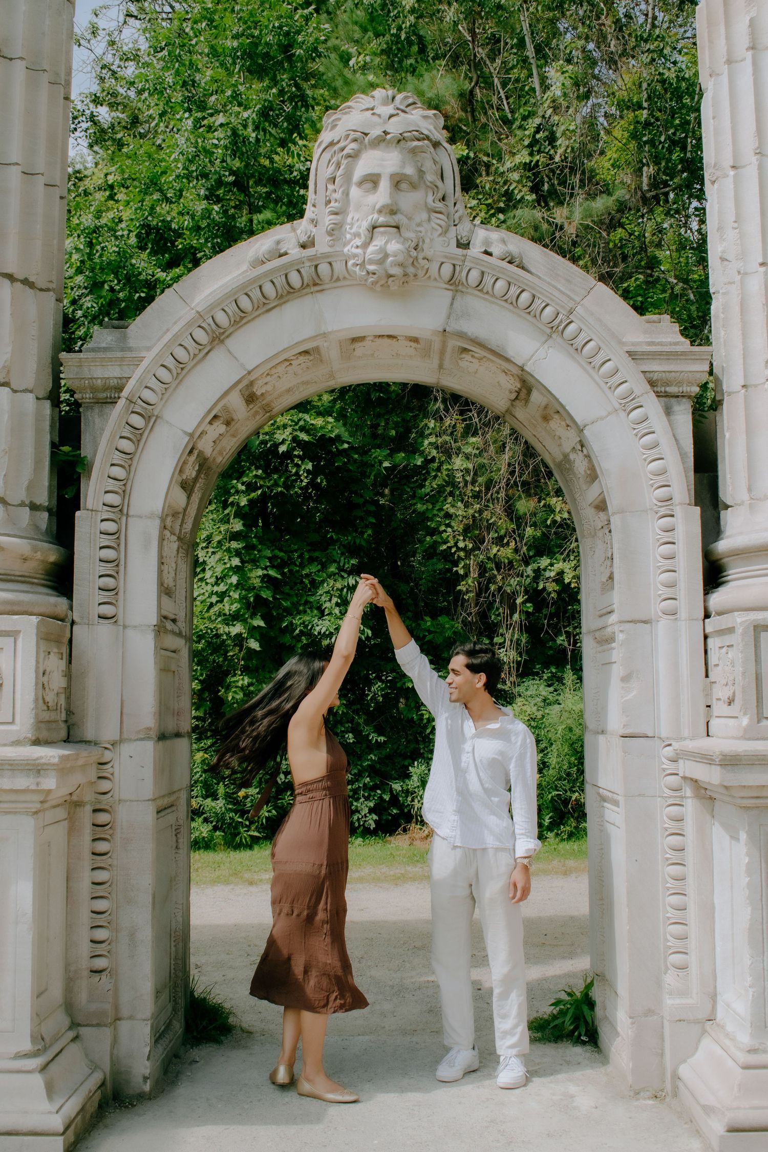 Couple dancing under ornate stone arch with classical face sculpture in lush green park setting.