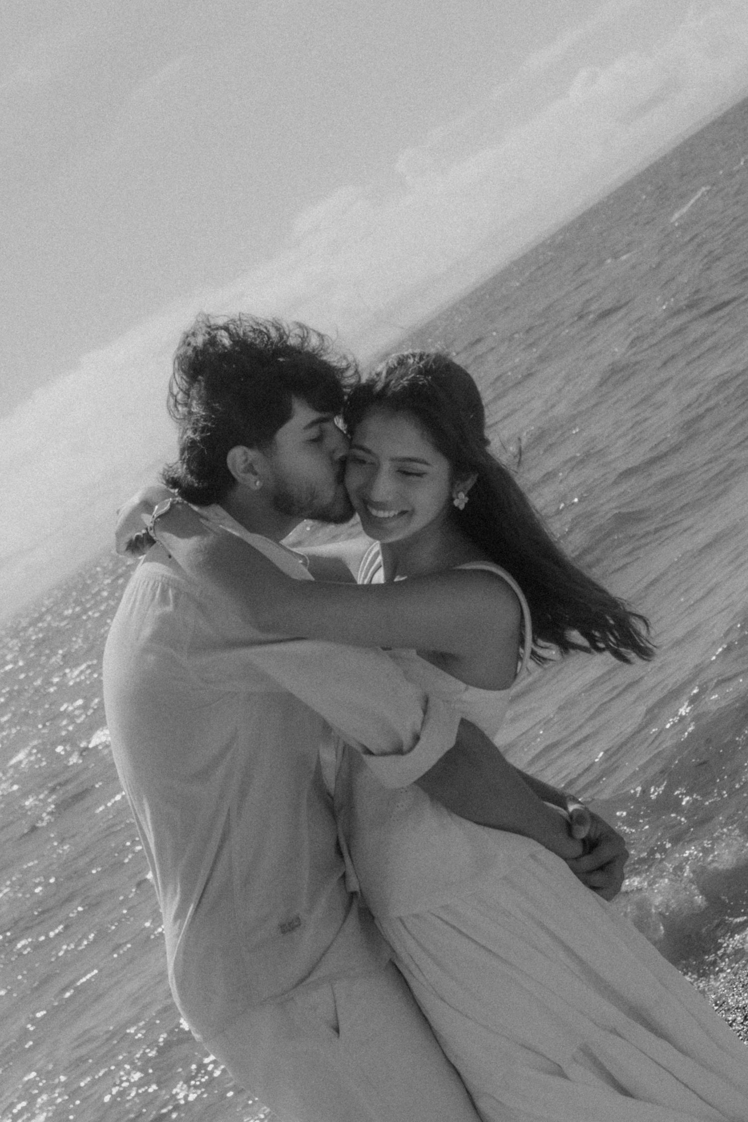 Black and white photo of a romantic couple embracing and kissing by the ocean shore.