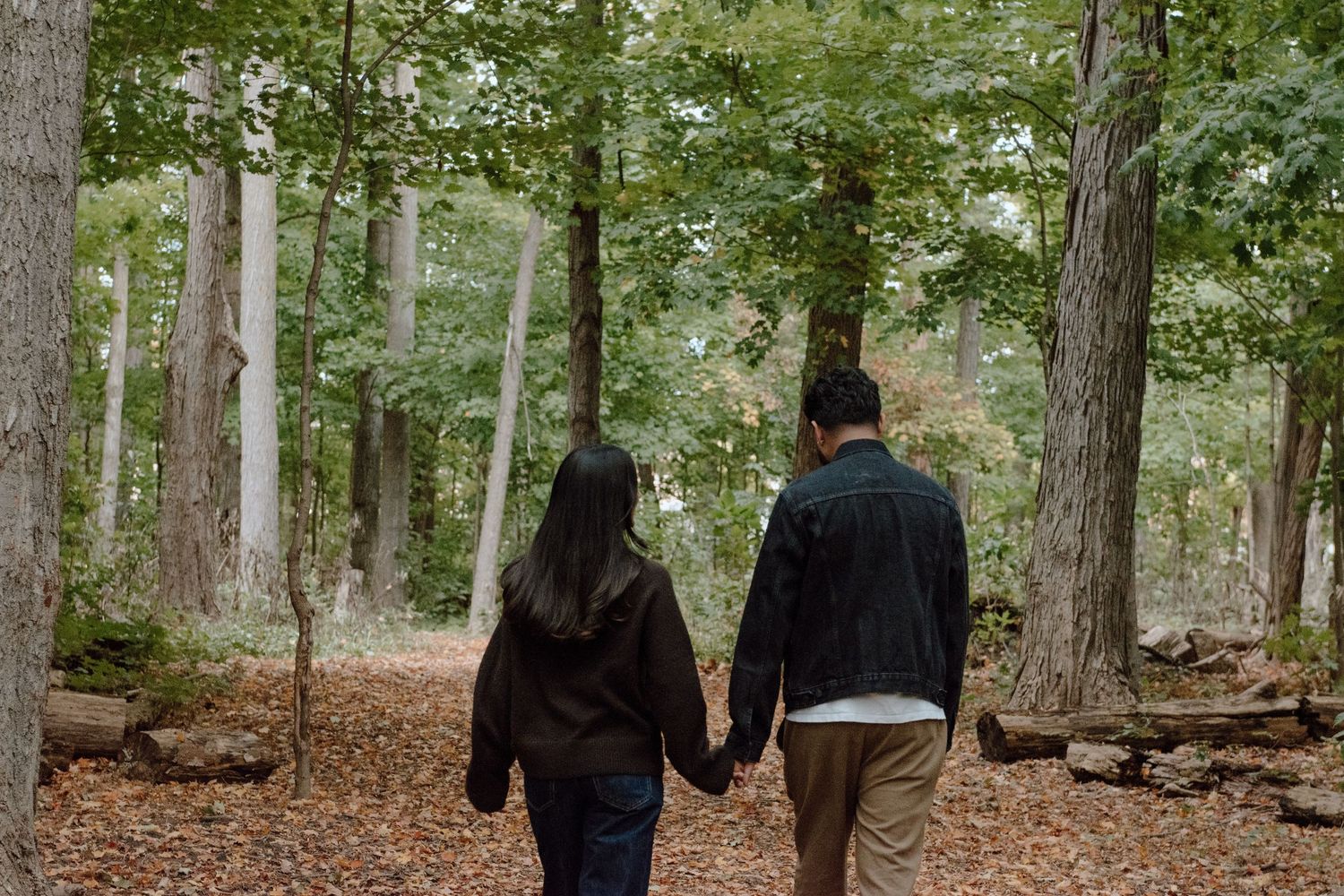 Couple holding hands walking through a forest trail covered in autumn leaves surrounded by tall trees.