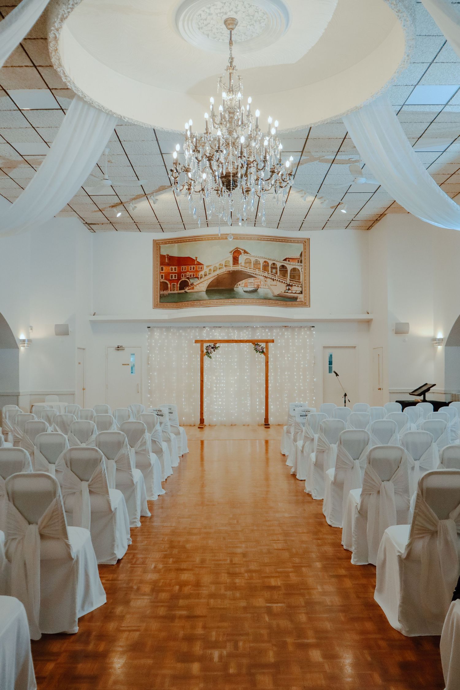 Elegant wedding ceremony hall with white chair covers, crystal chandelier, draped fabric ceiling, and a decorative arch at the altar.