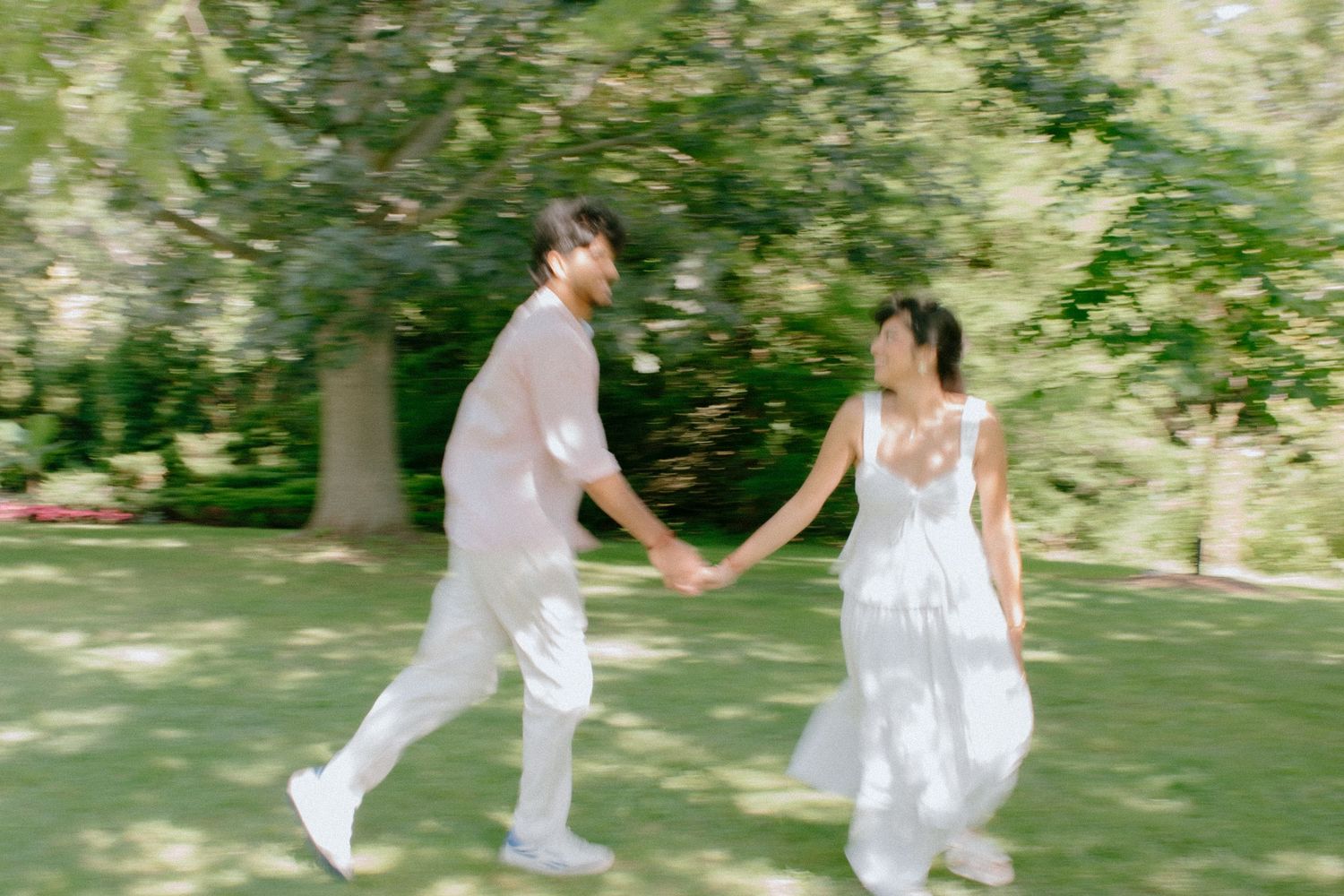 Couple in white outfits holding hands and walking through a lush green park on a sunny day.