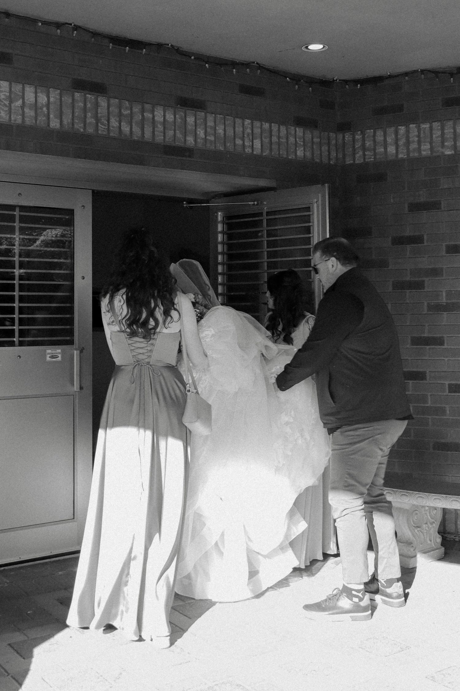 Black and white photo of bridesmaids helping bride with dress outside church.