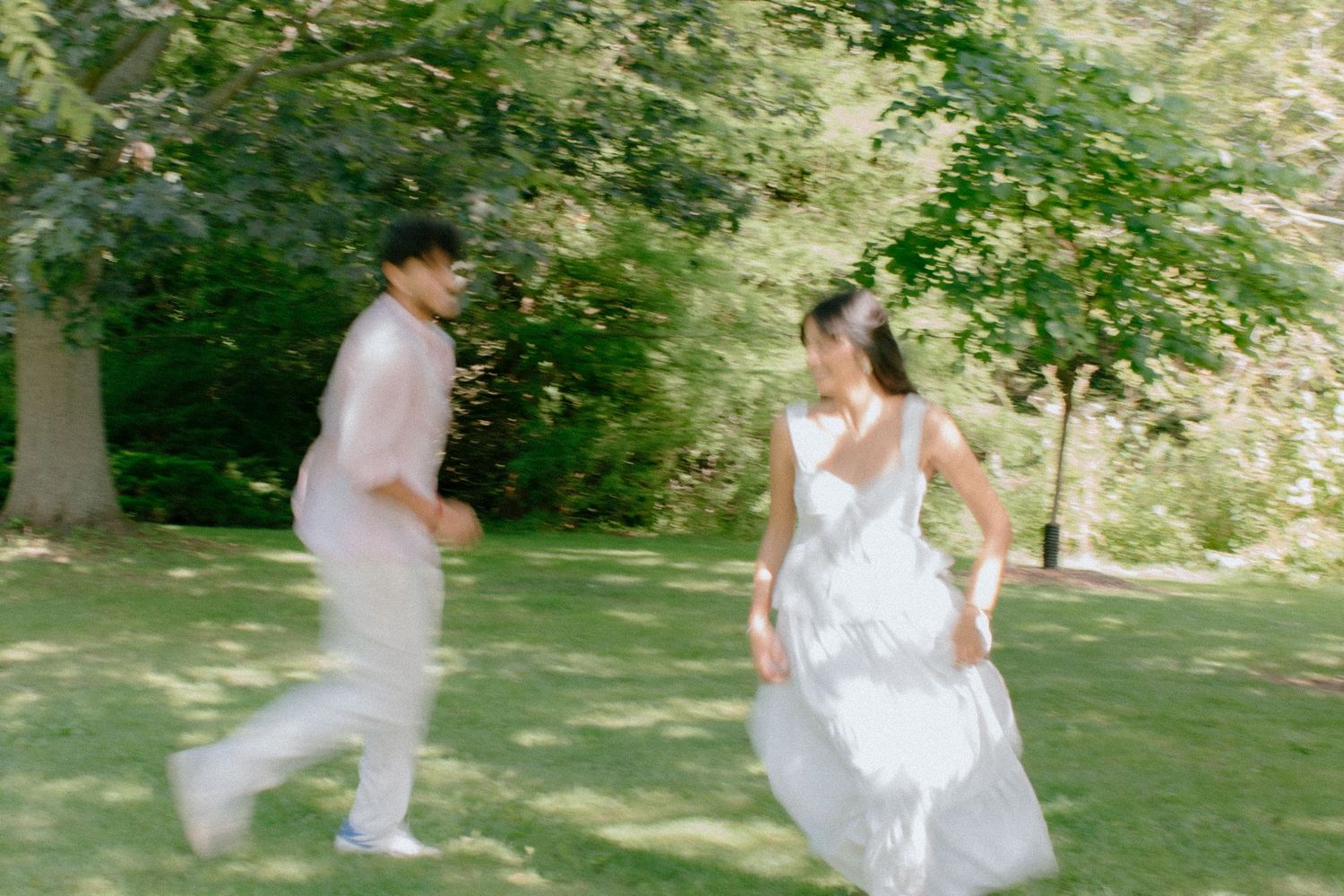 Couple in white attire running playfully through a lush green park on a sunny day.